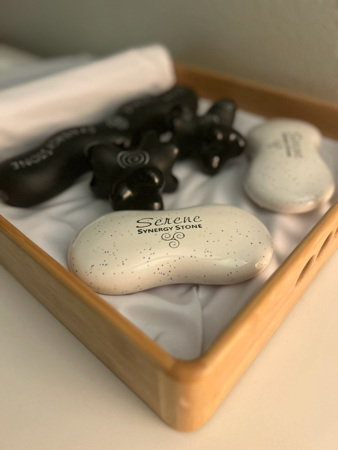 A gift box with a white silky cloth contains black massage stones and two white stones labeled 'Serene' and 'Synergy'.