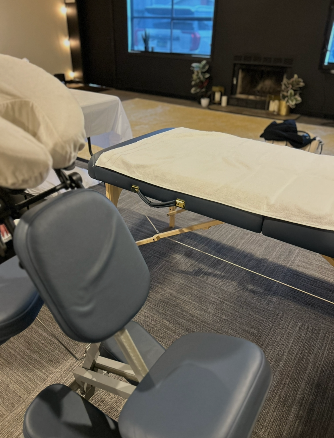 A massage table covered with a white towel, situated in a dimly lit room with a fireplace, candles, and decorative plants in the background.