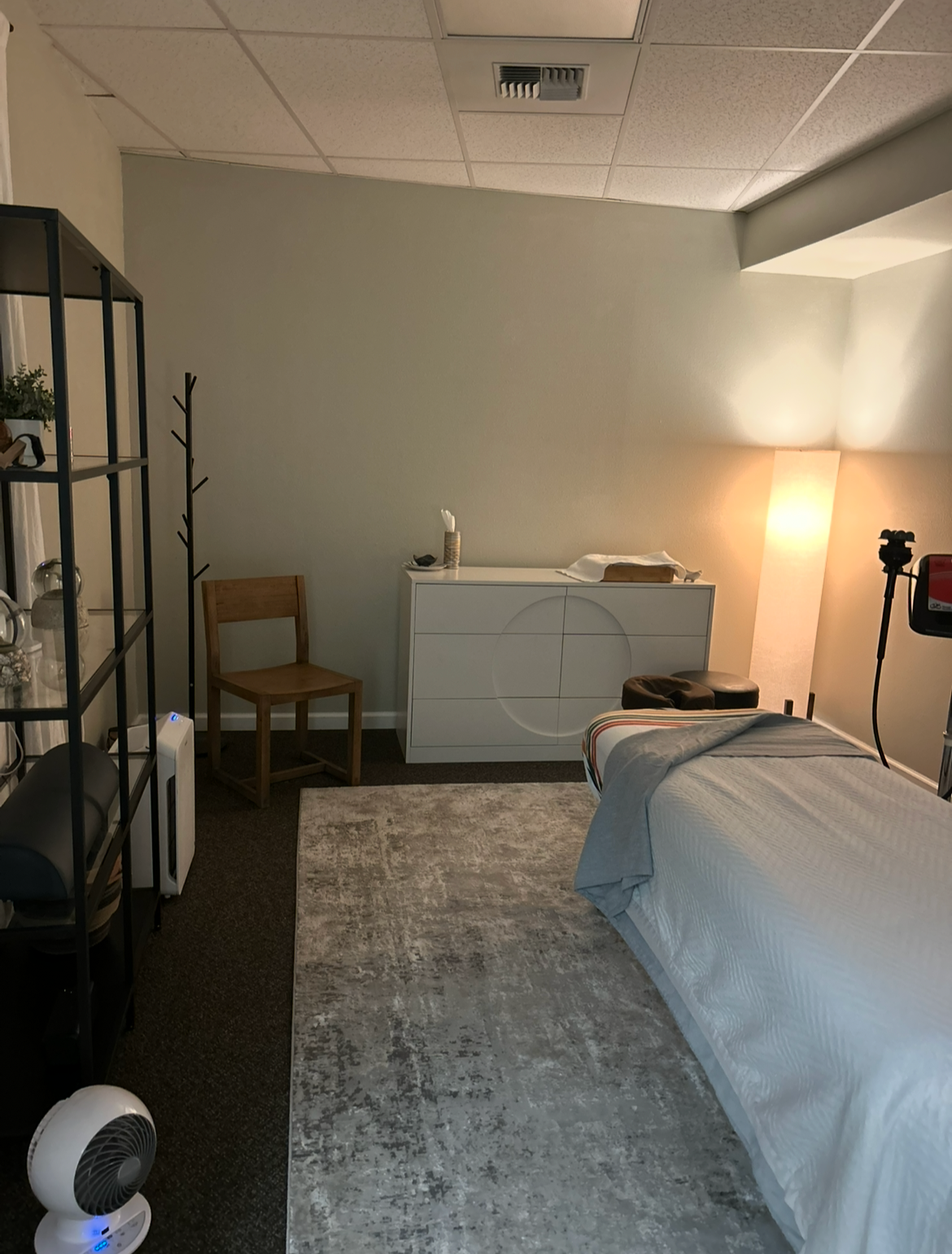 A cozy room with a bed, a wooden chair, a white dresser, a standing lamp, a coat rack, and a shelf with decorative items and plants.