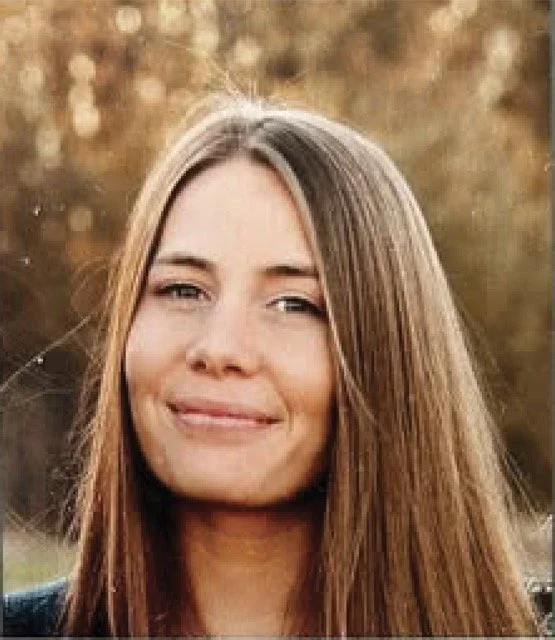 A young woman with long brown hair smiling outdoors, blurred autumn foliage background.