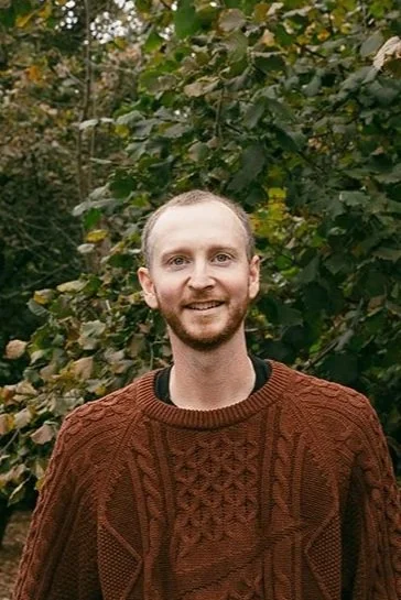 A man with light skin, short light hair, and a beard, standing outdoors in front of green leafy trees, wearing a brown patterned sweater.