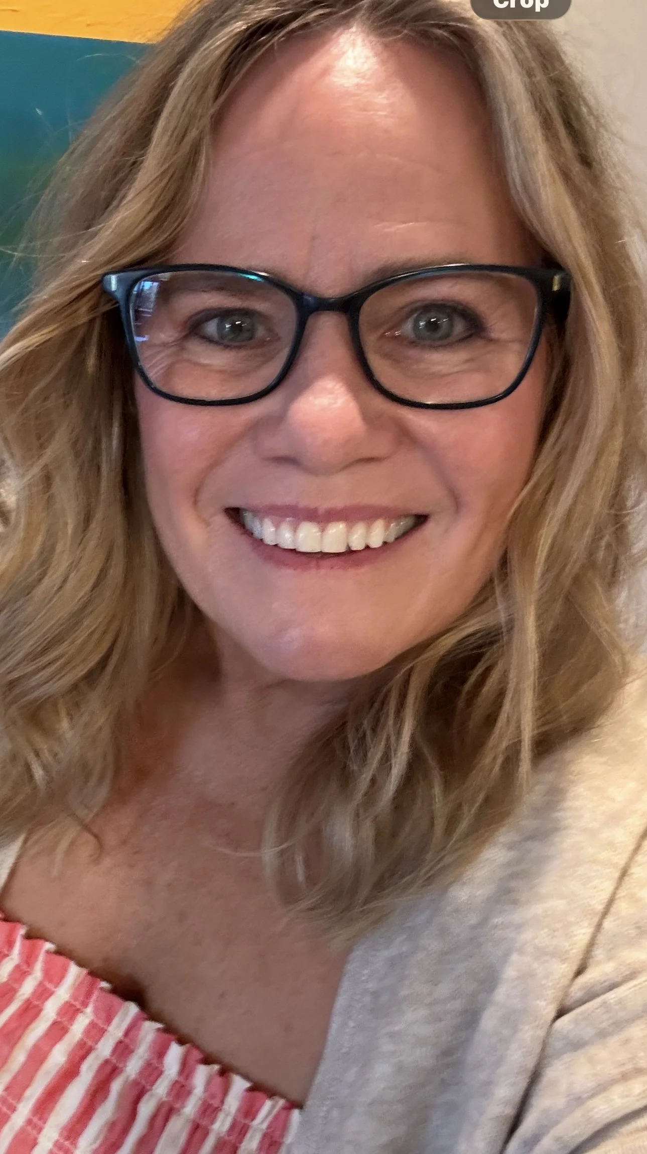 A smiling woman with long blonde wavy hair, glasses, and wearing a light-colored top.