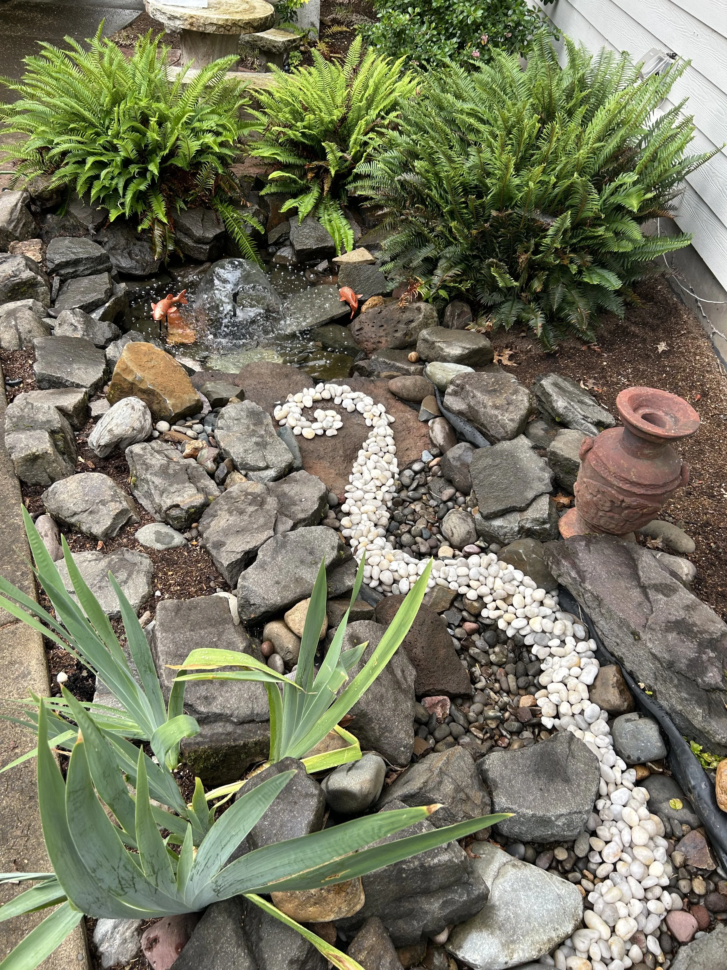 A small garden scene featuring lush green ferns behind a tiny bubbling water fountain, surrounded by rocks and decorative pebbles, with an assortment of plants and garden ornaments including a large ceramic vase and a barrel-shaped pot.