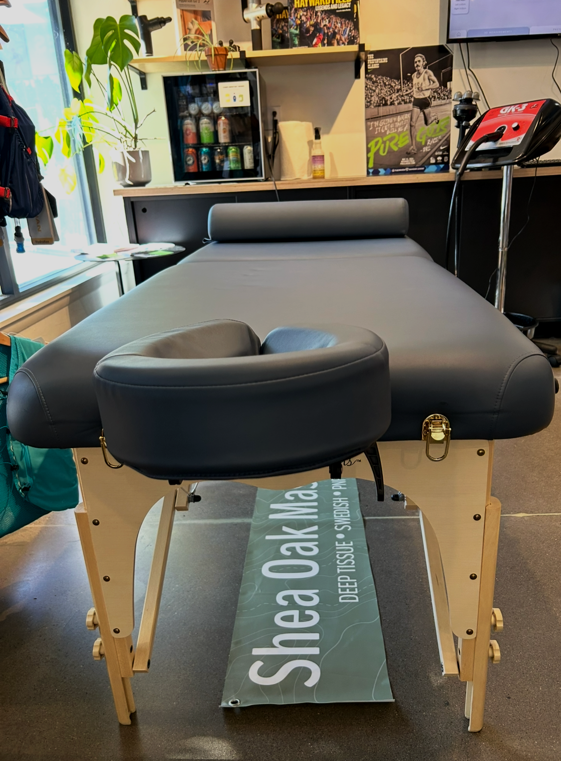 A massage table with a dark gray cushion and a face cradle, set up in a room with shelves, plants, posters, and a small refrigerator.