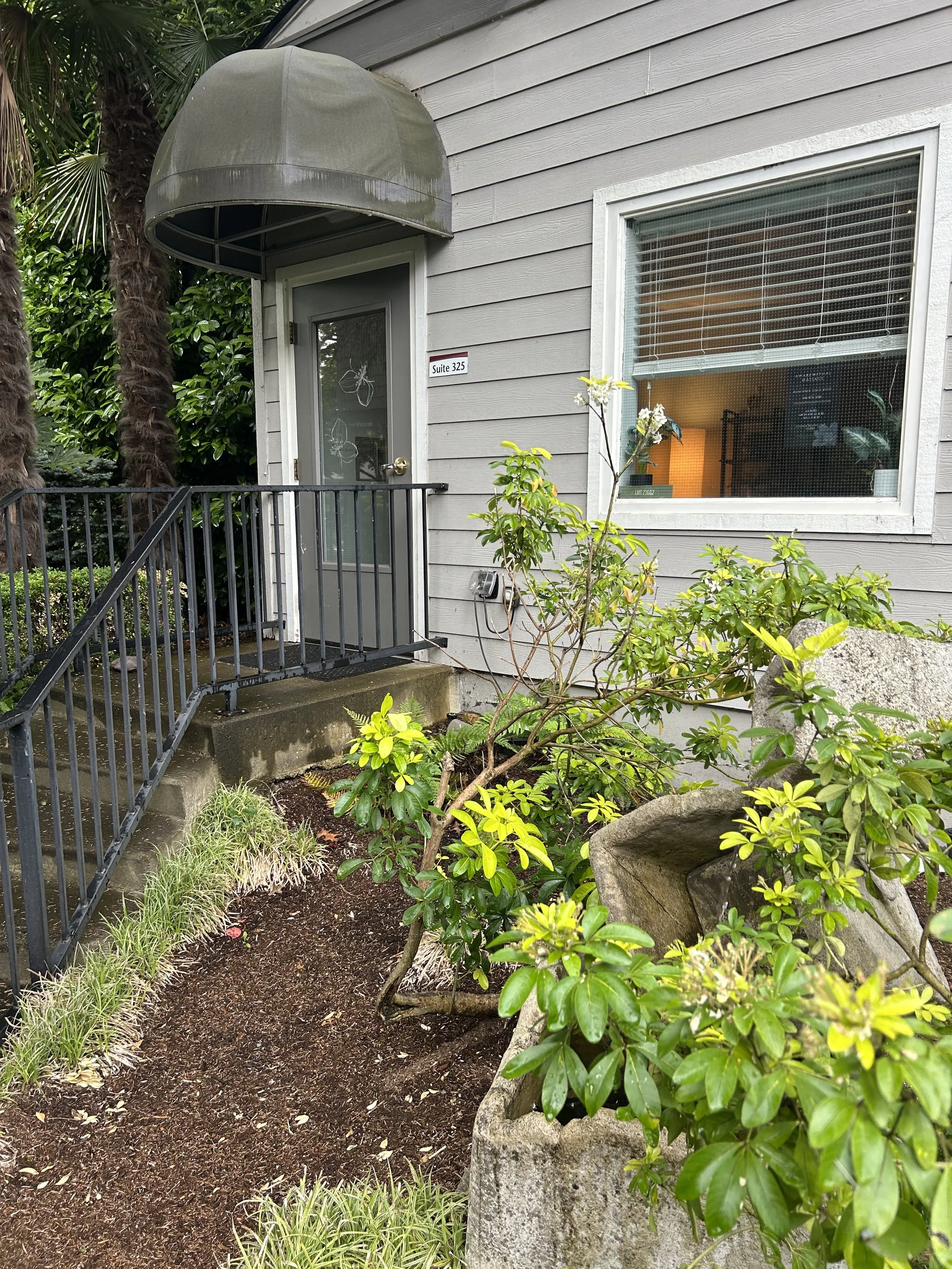 A residential entrance with a gray door with a glass panel, a black metal railing, and a large window with blinds, surrounded by shrubs and a garden bed, with a concrete step and a rock with greenery in the foreground.