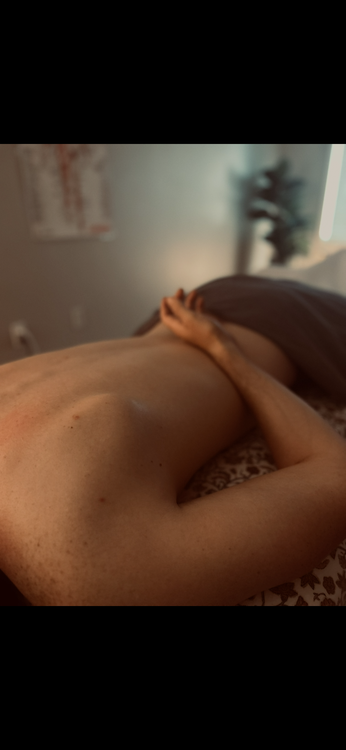 Person lying down on a patterned surface, with arm bent and hand resting on the side of their body. The background includes a wall with a poster and a blurred plant.