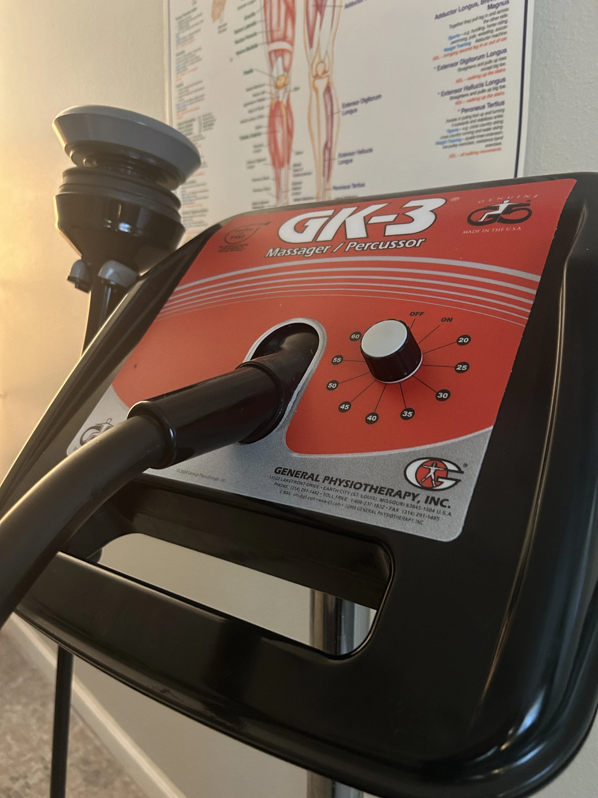 A massage machine with a dial and a red label on top, with a poster of human muscular anatomy in the background.