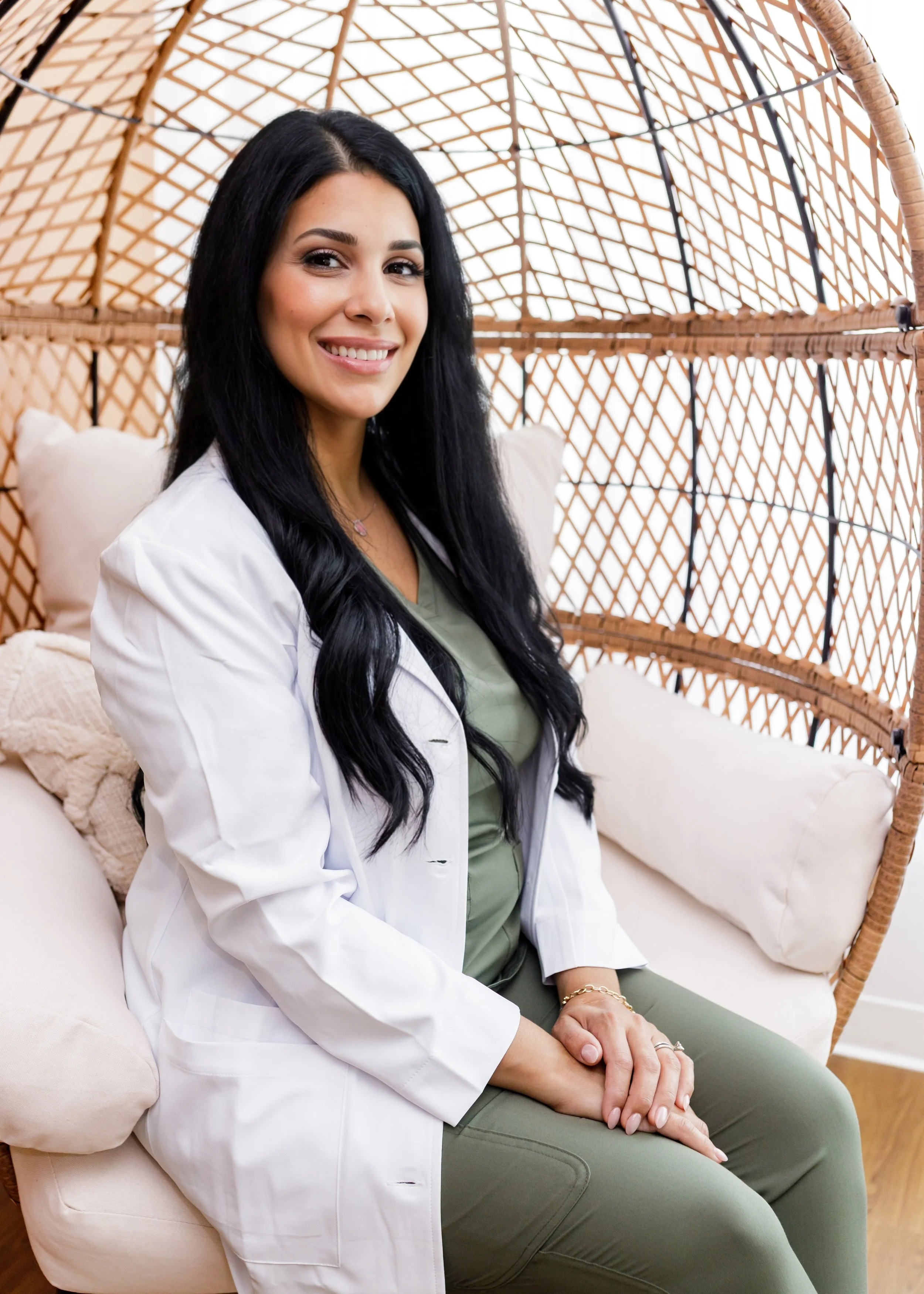 A woman with long black hair, wearing a white blazer and green scrubs, sitting on a beige cushioned sofa inside a wicker cocoon chair.