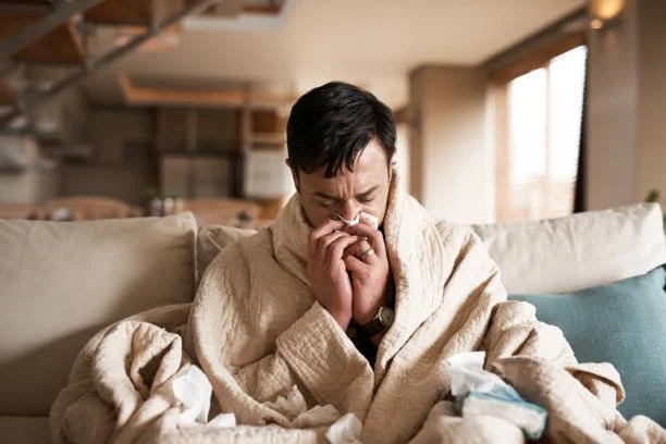 Man sitting on a couch with tissues, appearing to be sick or crying, in a cozy living room with sunlight from the window.