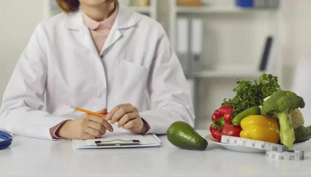 A person in a lab coat sitting at a table with fresh vegetables and a measuring tape.