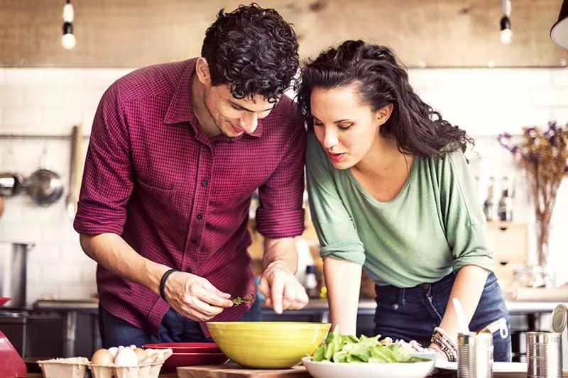 A man and woman preparing a meal together in a kitchen, leaning over a yellow bowl on the counter.