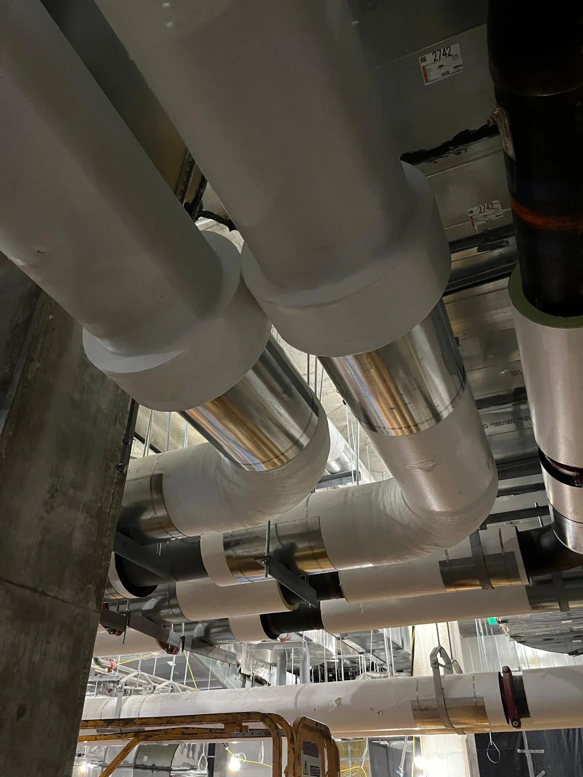 Large aluminum and white pipes installed on a building ceiling, secured with metal clamps, with some electrical wiring visible.