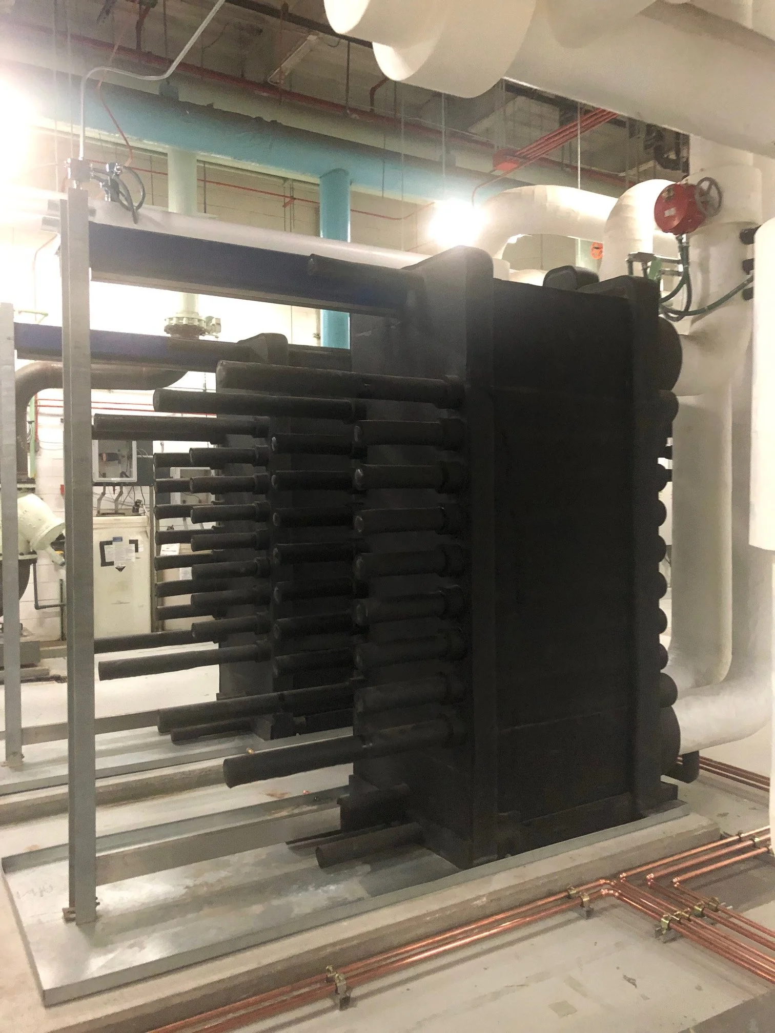 Industrial plumbing system including pipes, valves, and a large black equipment unit in a utility room.