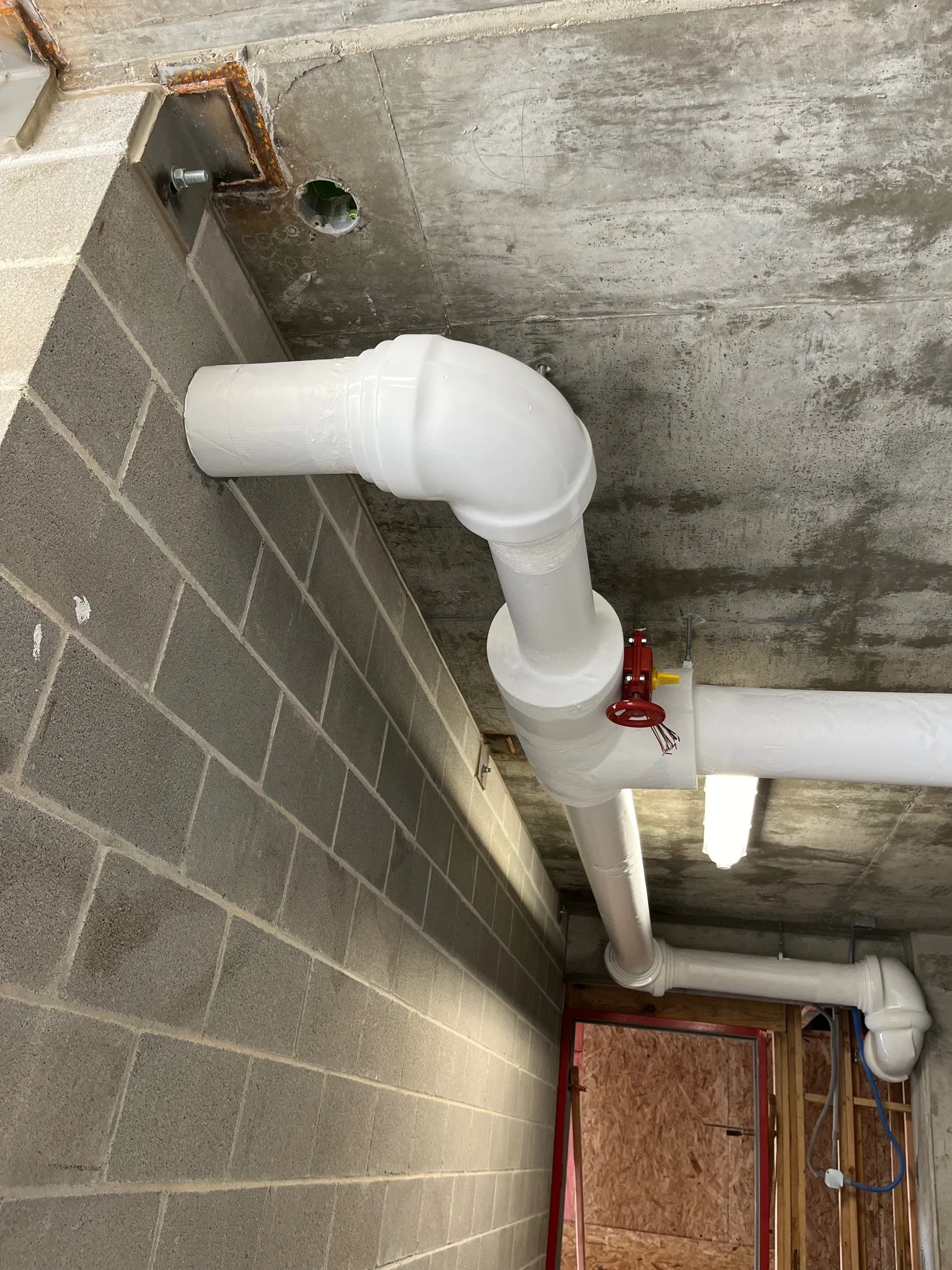 A construction area with exposed concrete walls, a white PVC pipe with a red valve, and unfinished framing and drywall in the background.