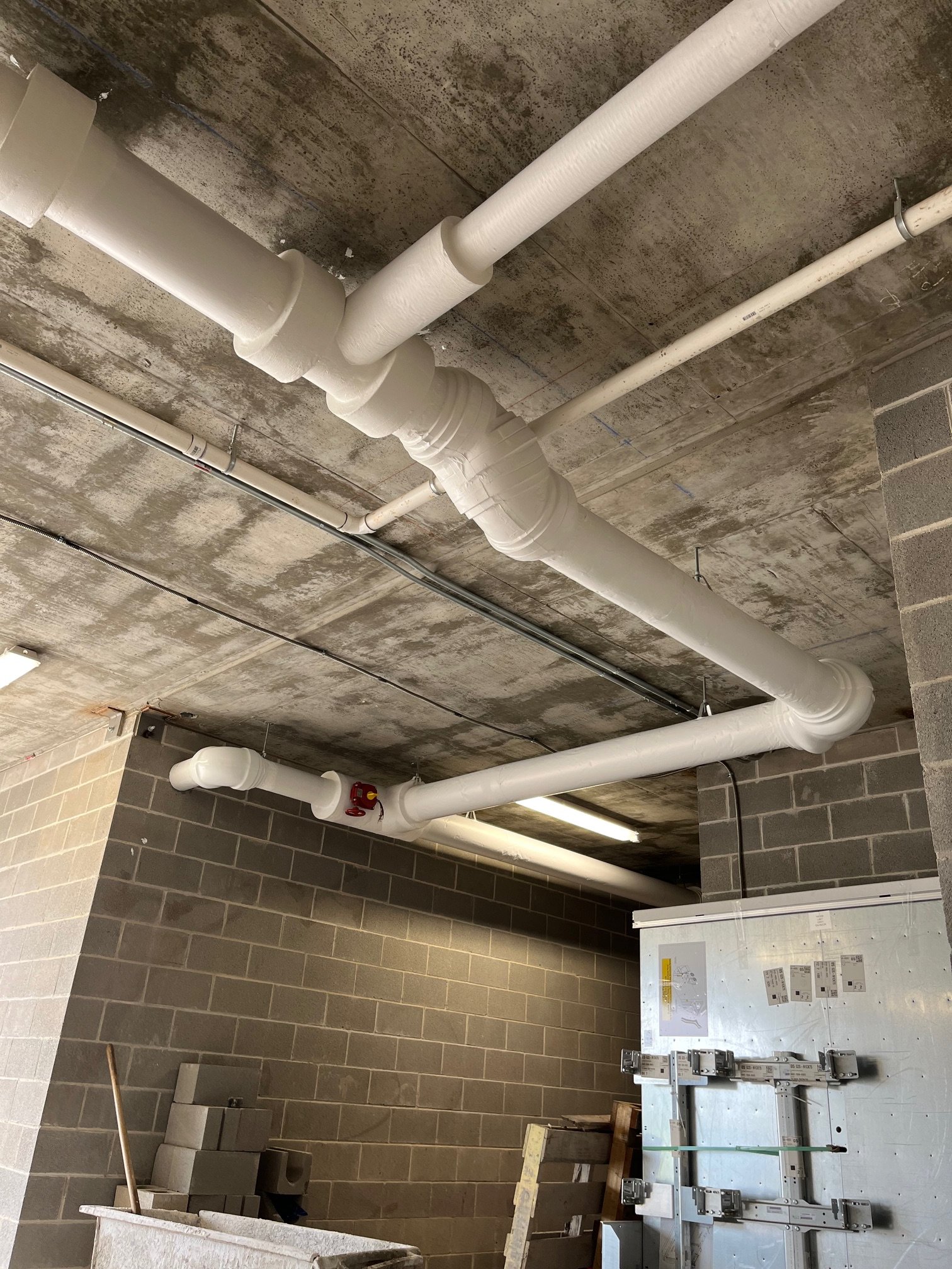 Construction area with gray cinder block walls and exposed concrete ceiling. White pipes run across the ceiling, some are large and curved. There is a white electrical cabinet with circuit breakers on the wall. Construction materials, equipment, and tools are scattered around.
