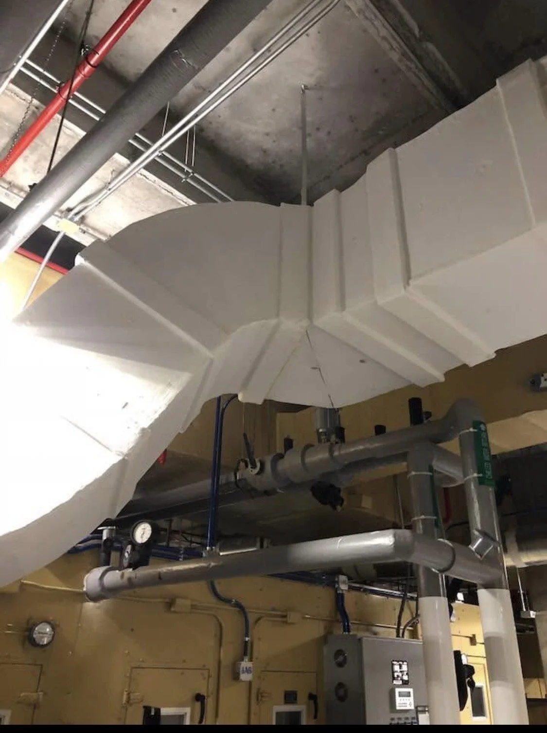 A large white ventilation duct installed on a ceiling with various pipes and electrical conduits nearby.