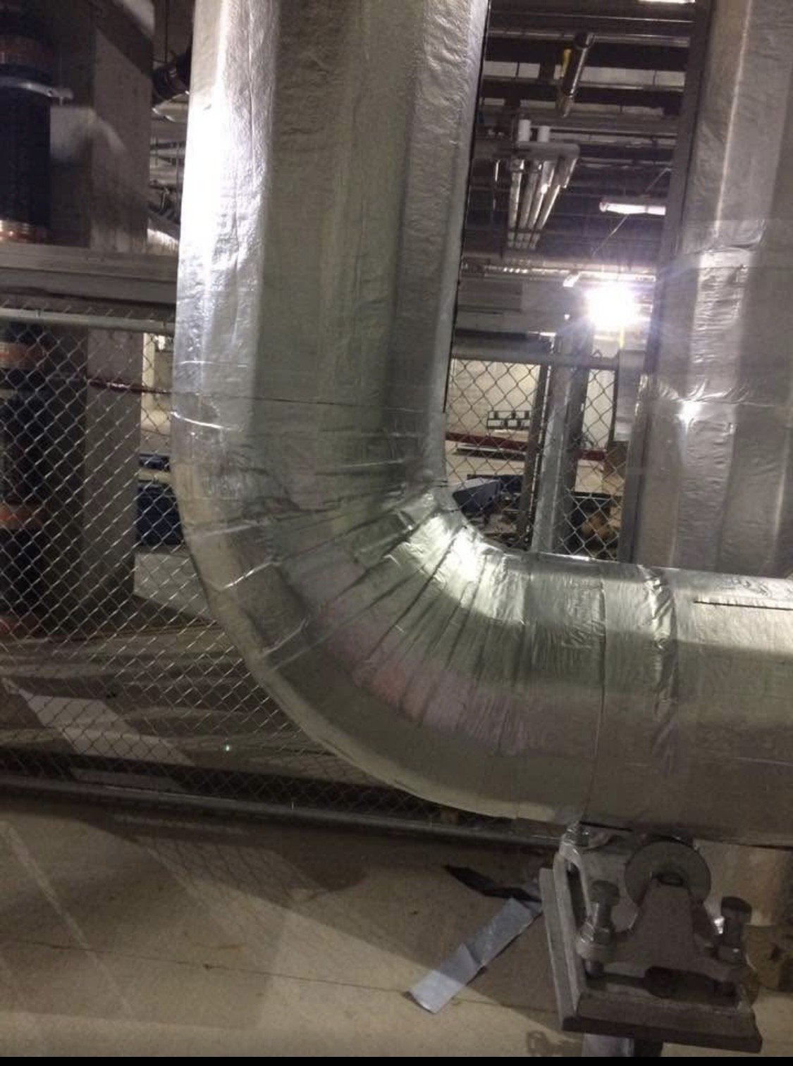 Large metallic ductwork with a curved section in an industrial or mechanical setting.