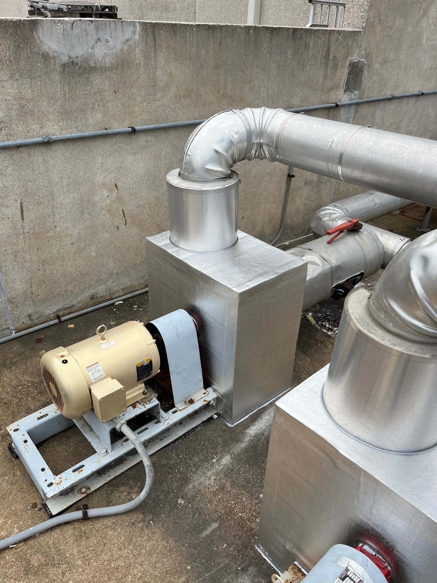Industrial rooftop machinery including silver pipes, a motor, and venting equipment mounted on a concrete surface with a wall in the background.