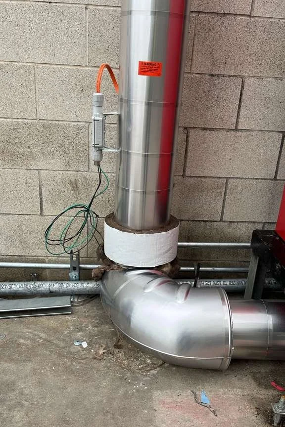 Industrial ductwork and piping, with a warning label on the tall vertical pipe, attached to a wall in a utility or mechanical room.