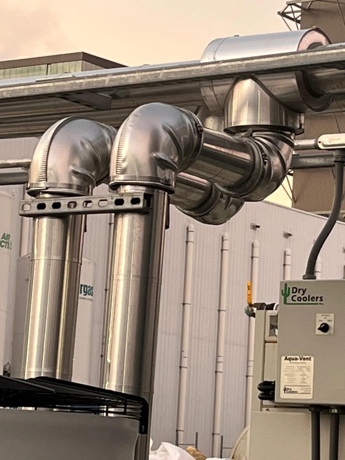 Industrial piping system with stainless steel pipes and a control unit labeled 'Dry Coolers' on a rooftop or industrial building.