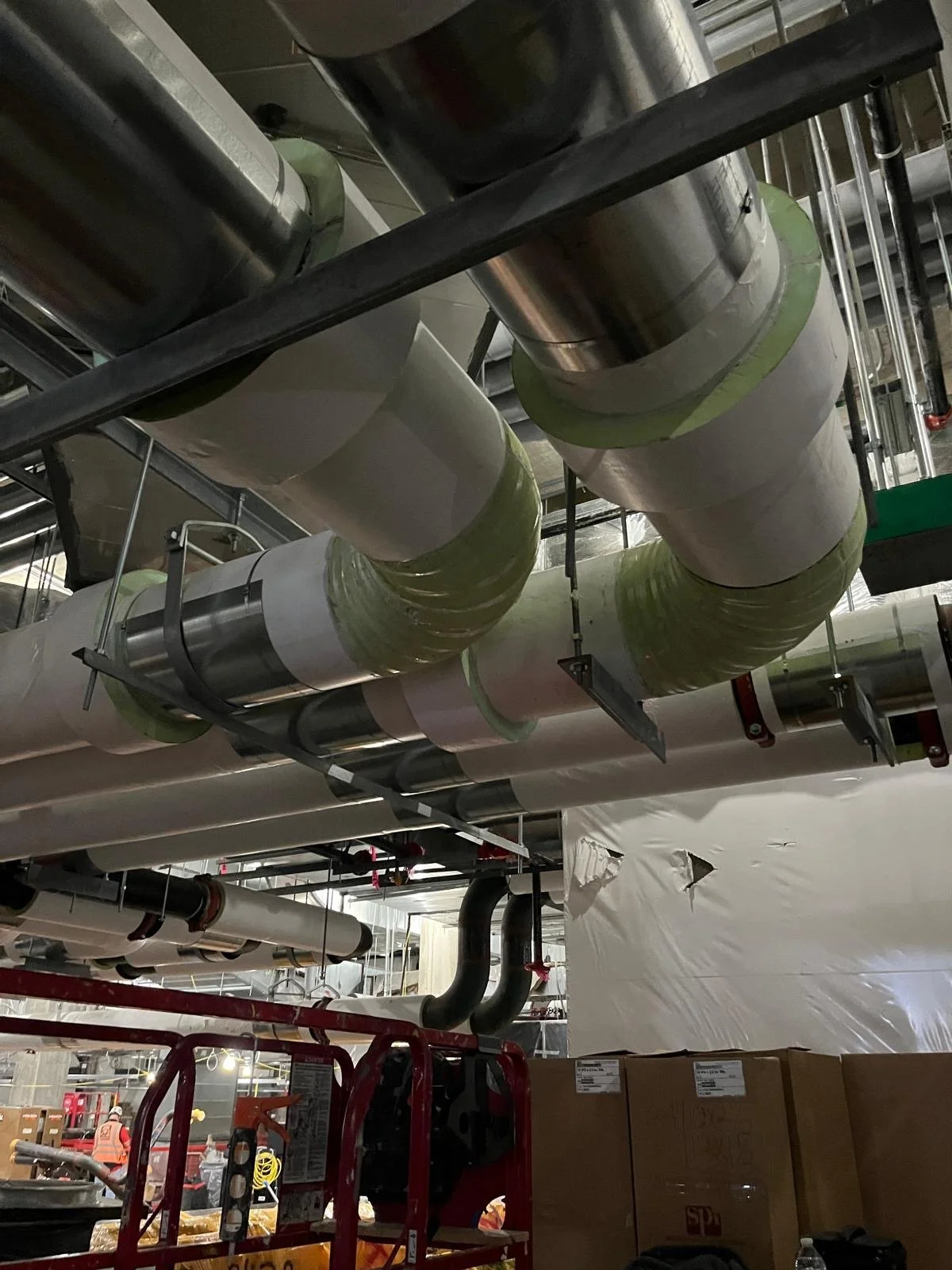 Ceiling pipes in an industrial or mechanical room with workers and boxes visible below.