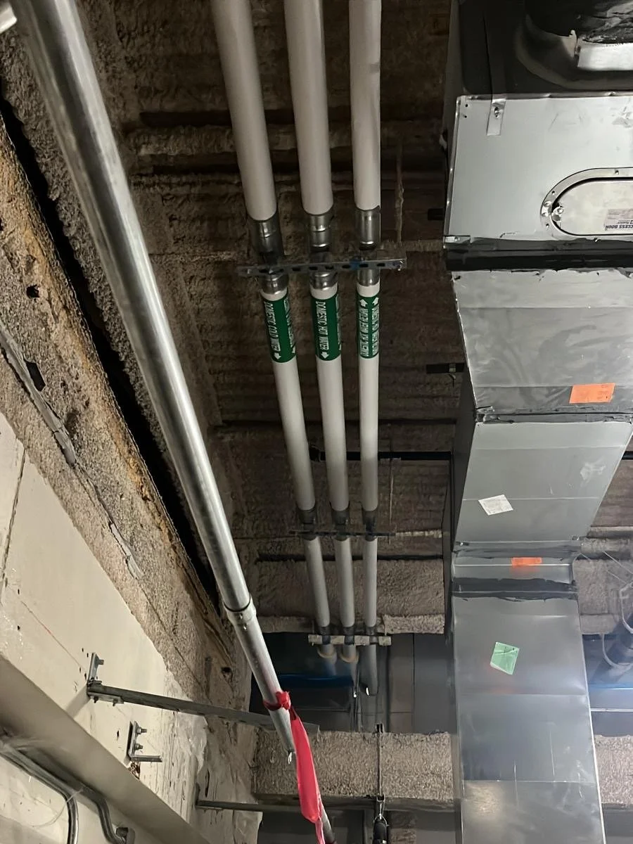 Ceiling view of a construction site showing white pipes, metallic ductwork, and electrical wiring installed on a ceiling with exposed brick and concrete.