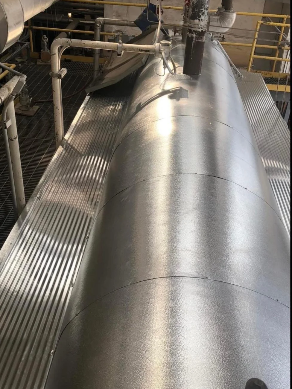 Close-up of a large, shiny metallic industrial pipeline or vessel in a factory setting with surrounding pipes and safety railings.