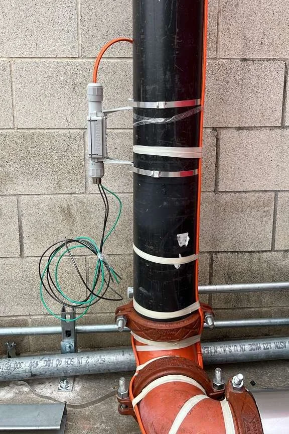 A large black plastic pipe connected to an orange pipe with white straps, with wiring attached to a white connector on the wall.