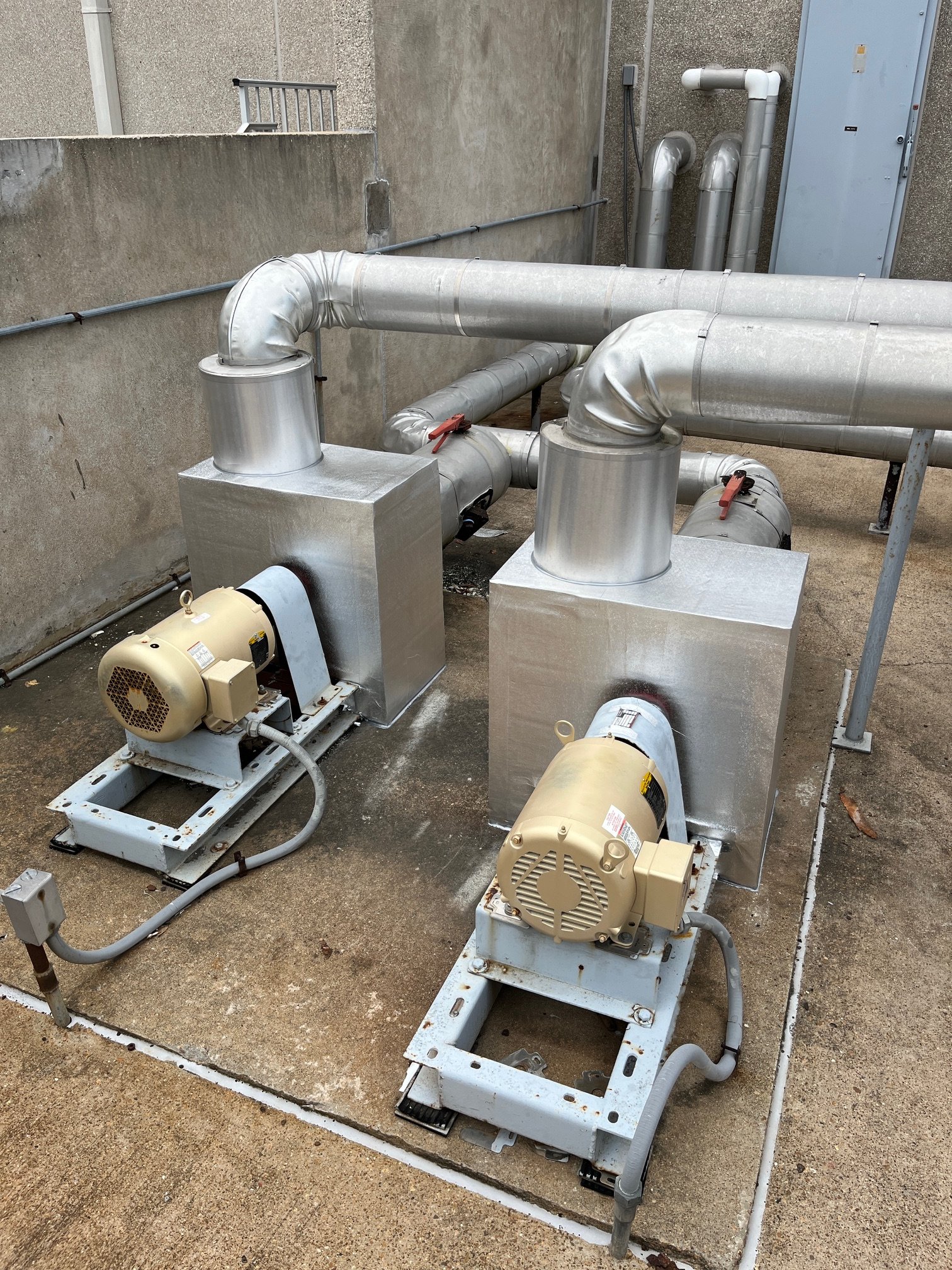 Two large pieces of industrial HVAC equipment with motors, mounted on metal frames on concrete ground, with duct pipes connected, set against a wall.