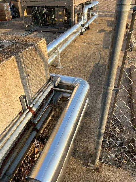 Silver industrial pipes running along a concrete surface, next to a chain-link fence and a concrete block, with machinery in the background.
