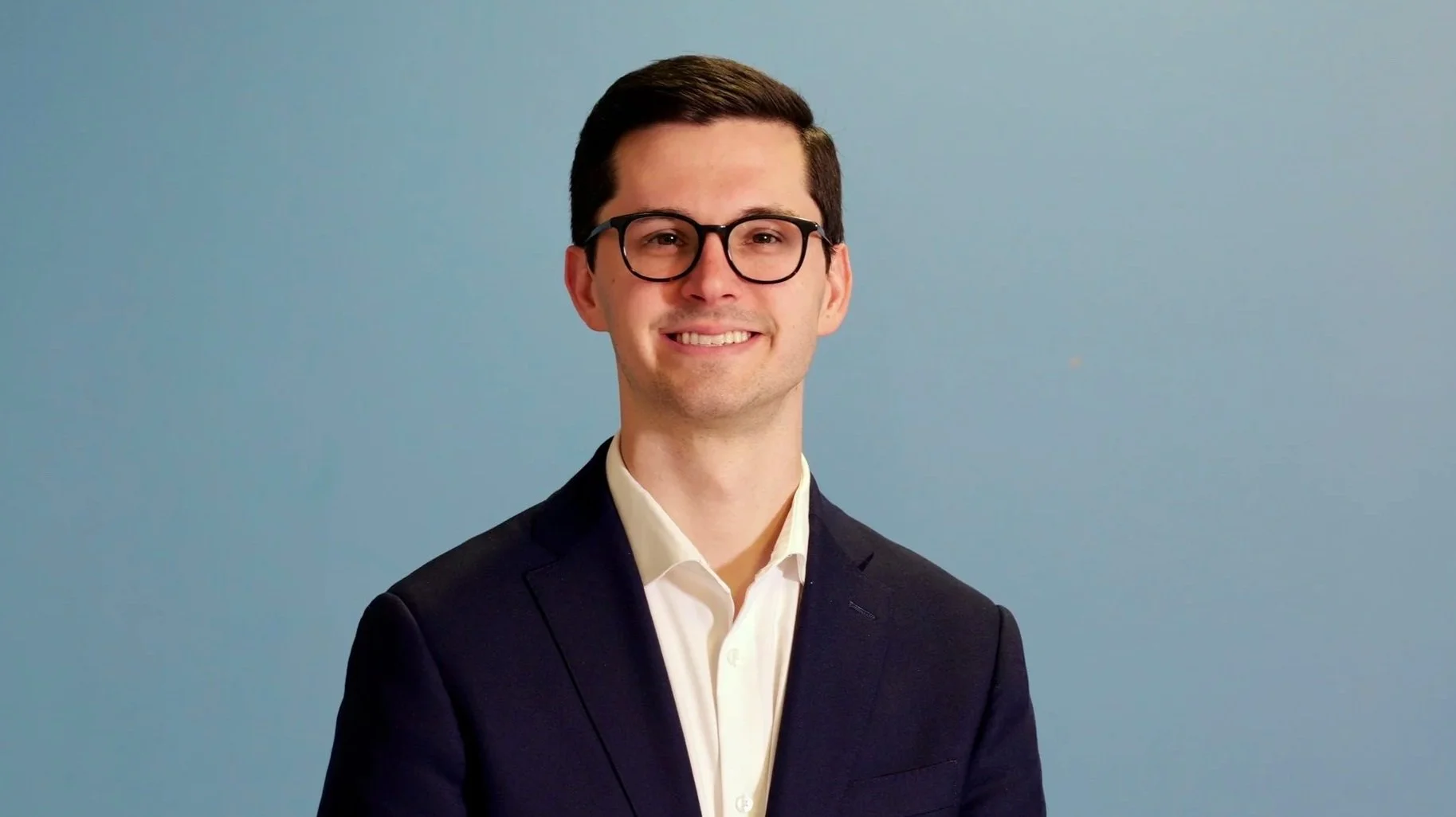 A portrait of a young man with dark hair, glasses, and a friendly smile, wearing a dark suit and white shirt, standing against a light blue background.
