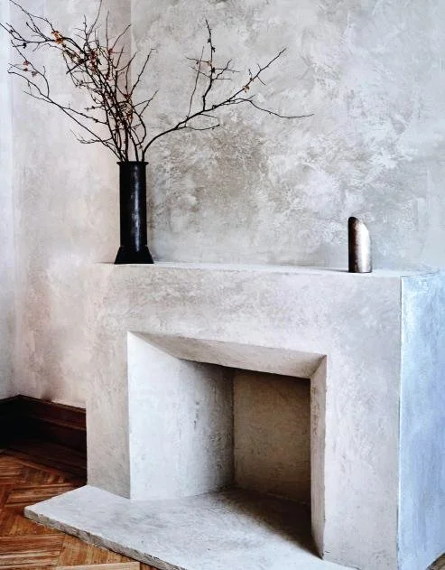 A minimalist fireplace with a black vase holding dry branches and a small rounded object on the mantle, against a textured light gray wall.