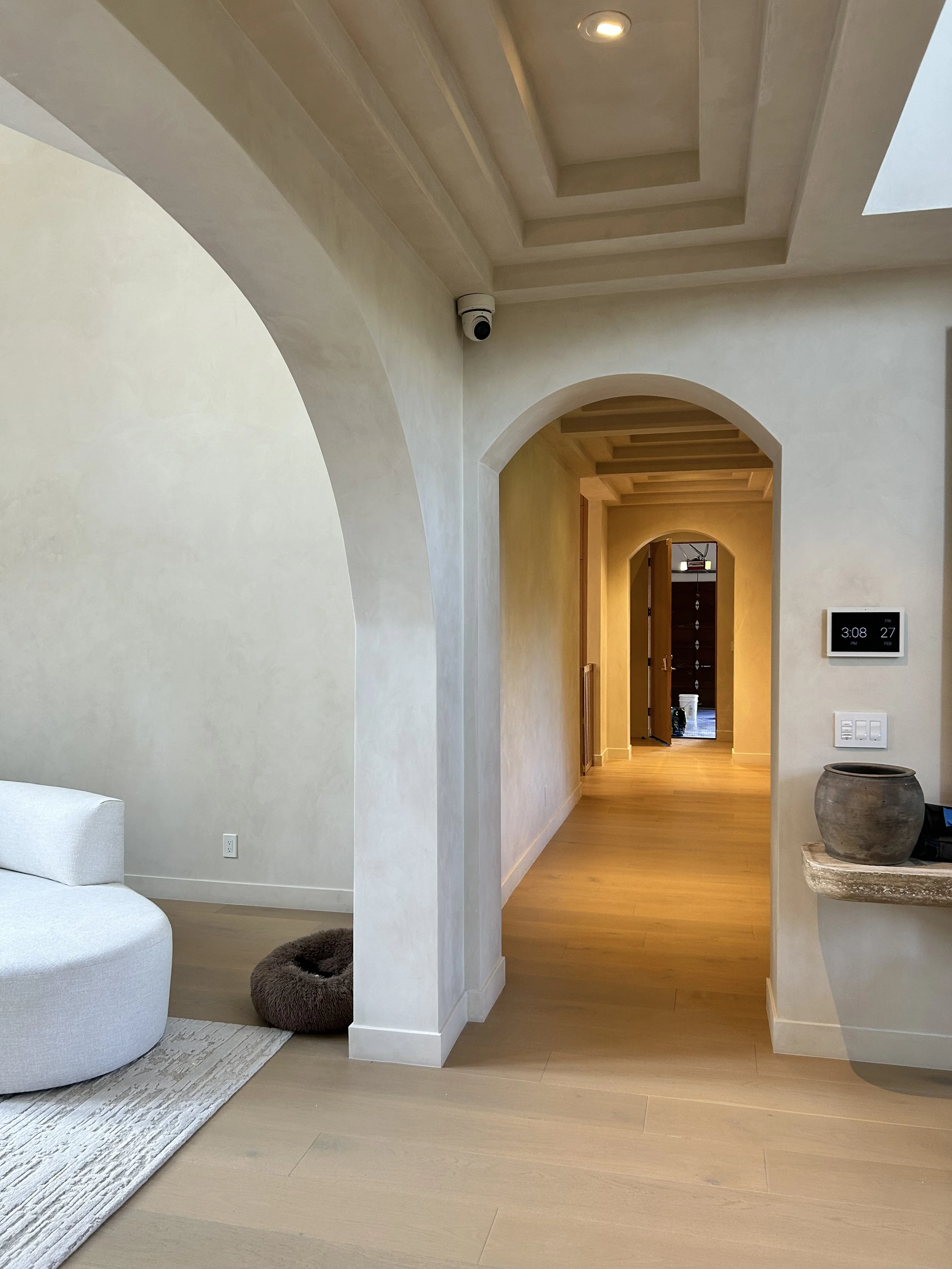 A hallway with wooden floors and beige walls, leading to another room through an arched doorway. The ceiling has recessed square designs with a t-shaped light. There is a security camera mounted in the corner, and a digital clock and light switches a