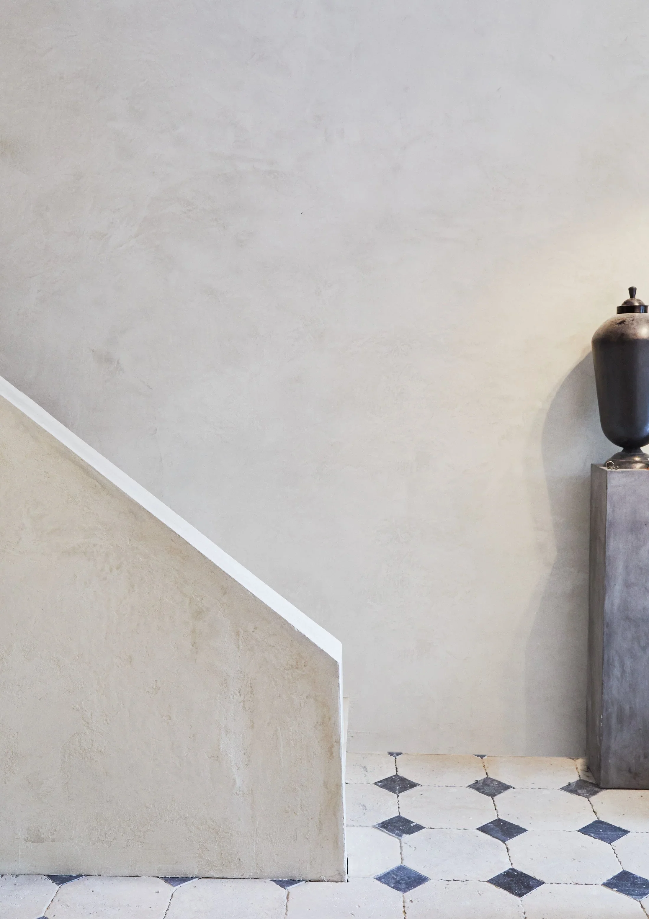 A beige wall with a textured finish, adjacent to a staircase with white steps and a handrail. On the right side, there is a large black urn on a stand, with a small sculpture or object on top.