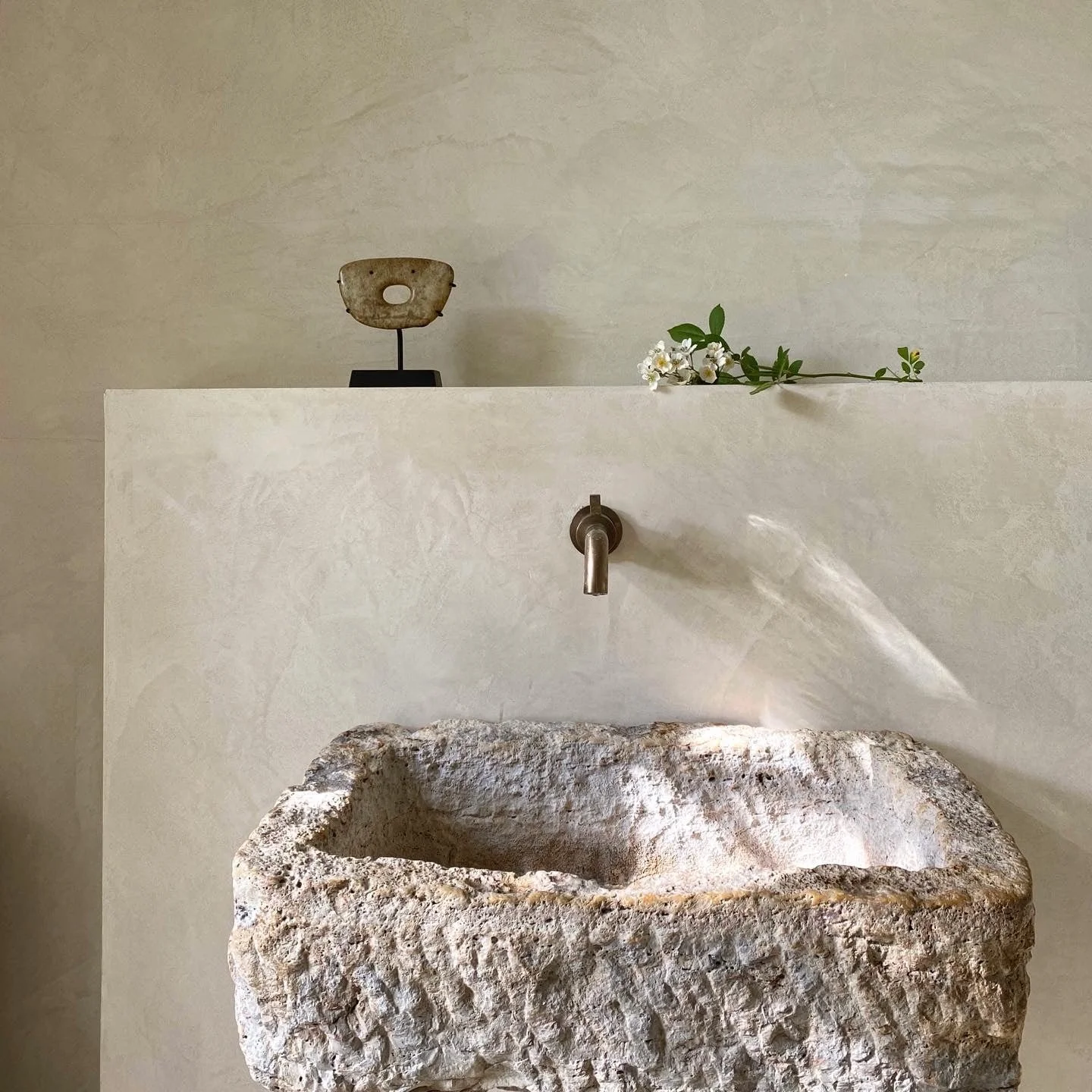 Stone sink with rough texture in a minimalist setting, featuring a modern faucet, placed against a neutral textured wall, with a black and white sculpture and a small flower arrangement on a surface above.