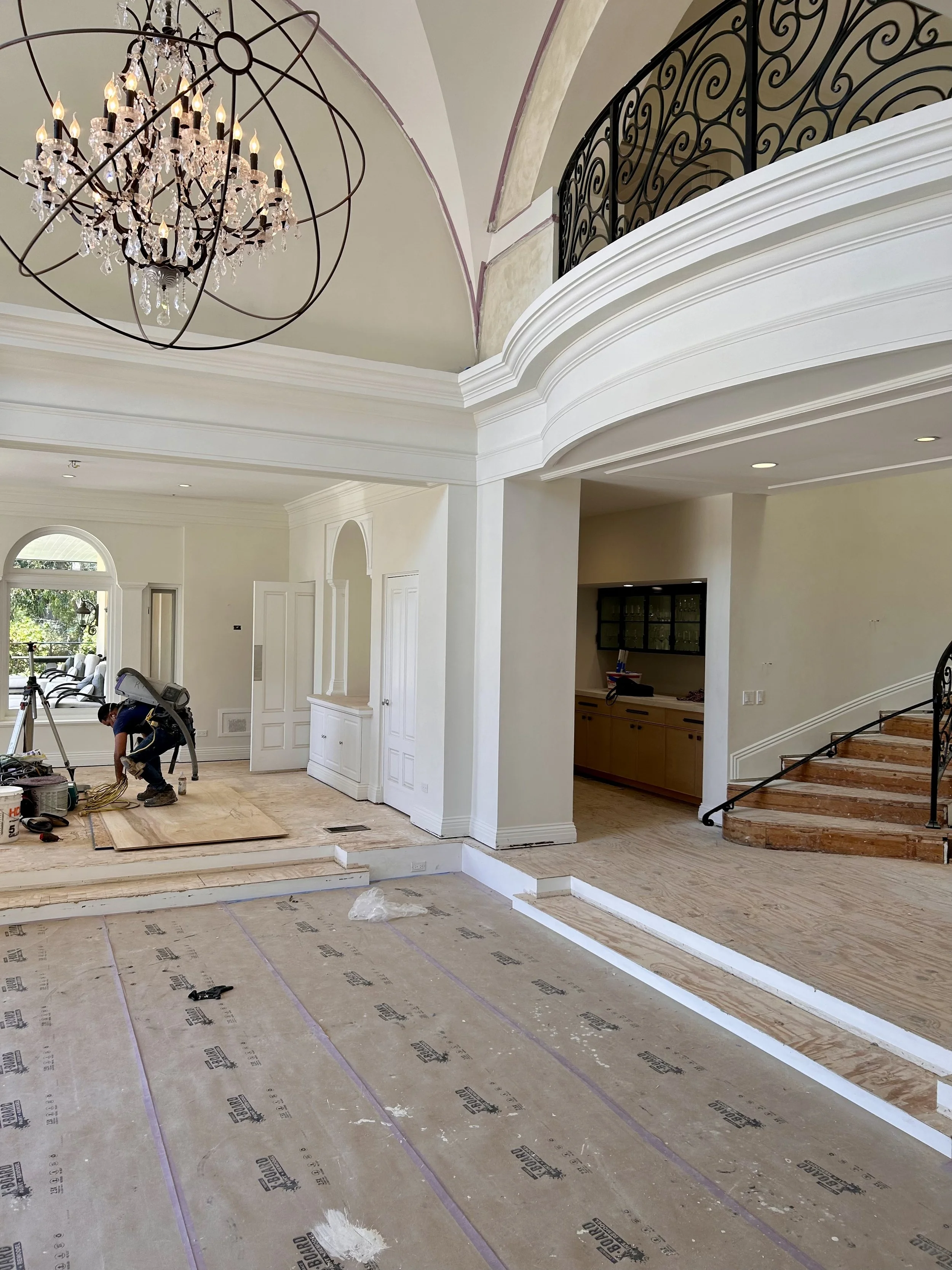 Interior of a house under renovation, with a worker installing hardwood flooring. The room features a chandelier hanging from the ceiling, a staircase with a wrought iron railing, and large windows.