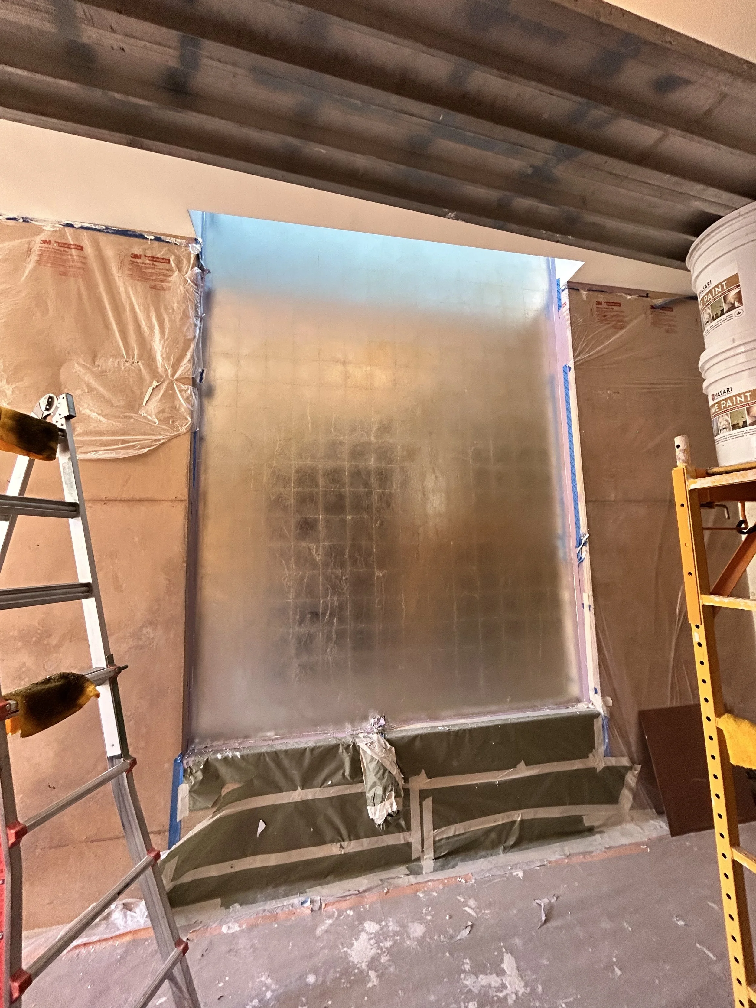 Interior construction site showing a partially framed wall, insulation wrapped in plastic, a ladder, and buckets of paint, with a window opening in the background.