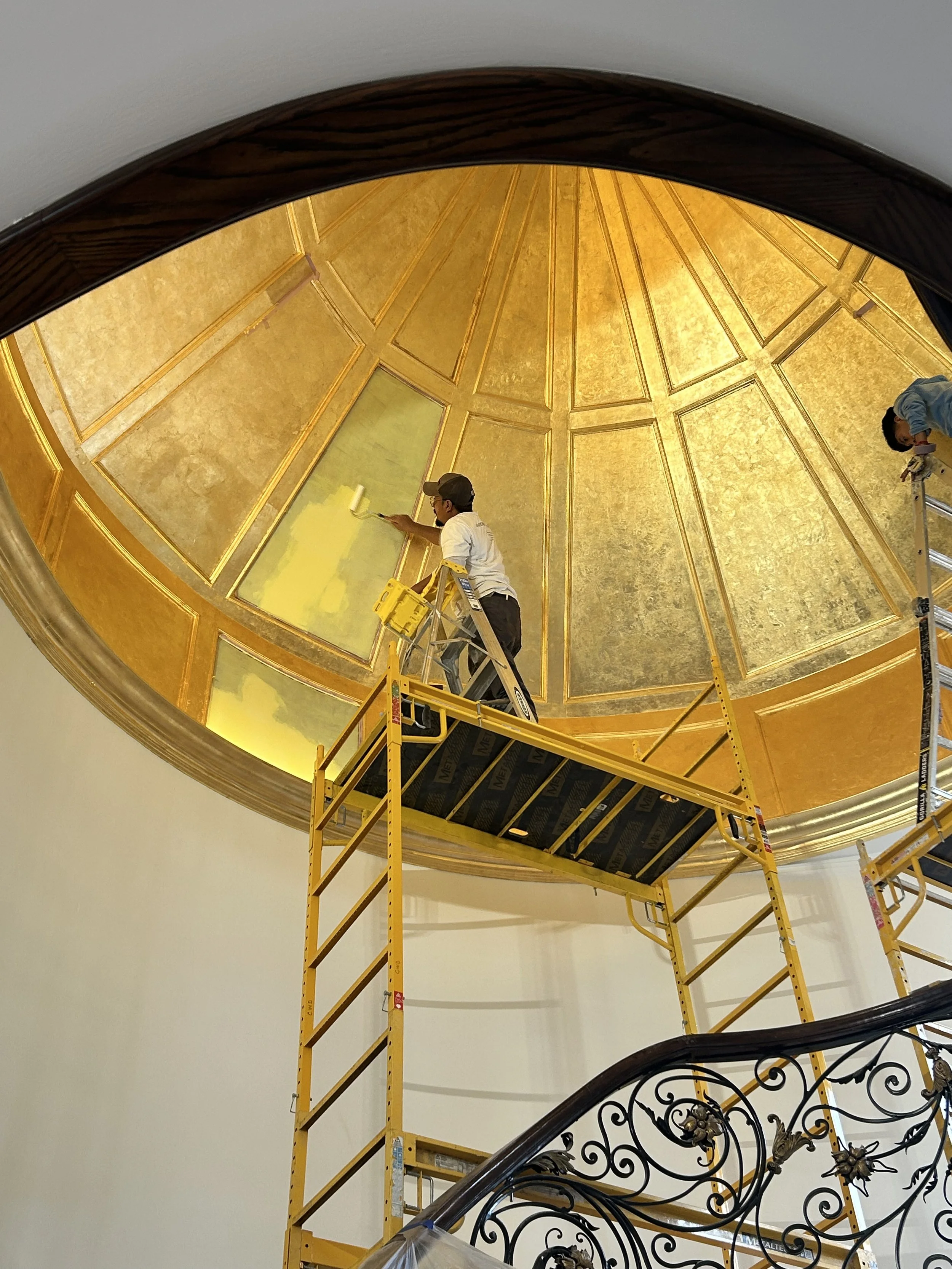 Workers standing on a yellow scaffold cleaning and restoring a gold-leafed dome ceiling with decorative paneling in an elegant building.