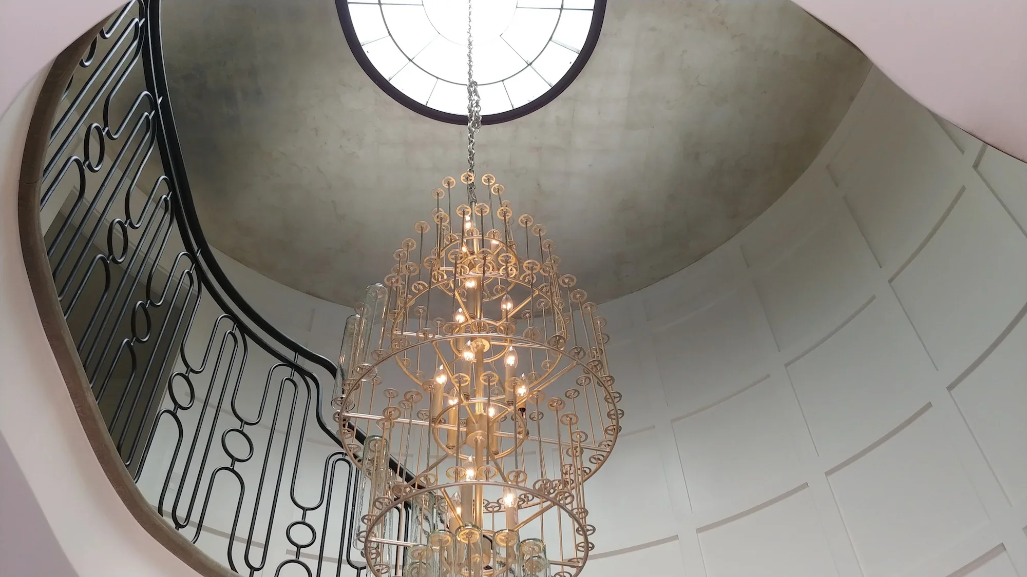 An elegant chandelier hanging from the ceiling near a circular skylight. The chandelier features a gold framework with numerous small lights and glass rods, and the ceiling is painted in a soft hue with architectural details.