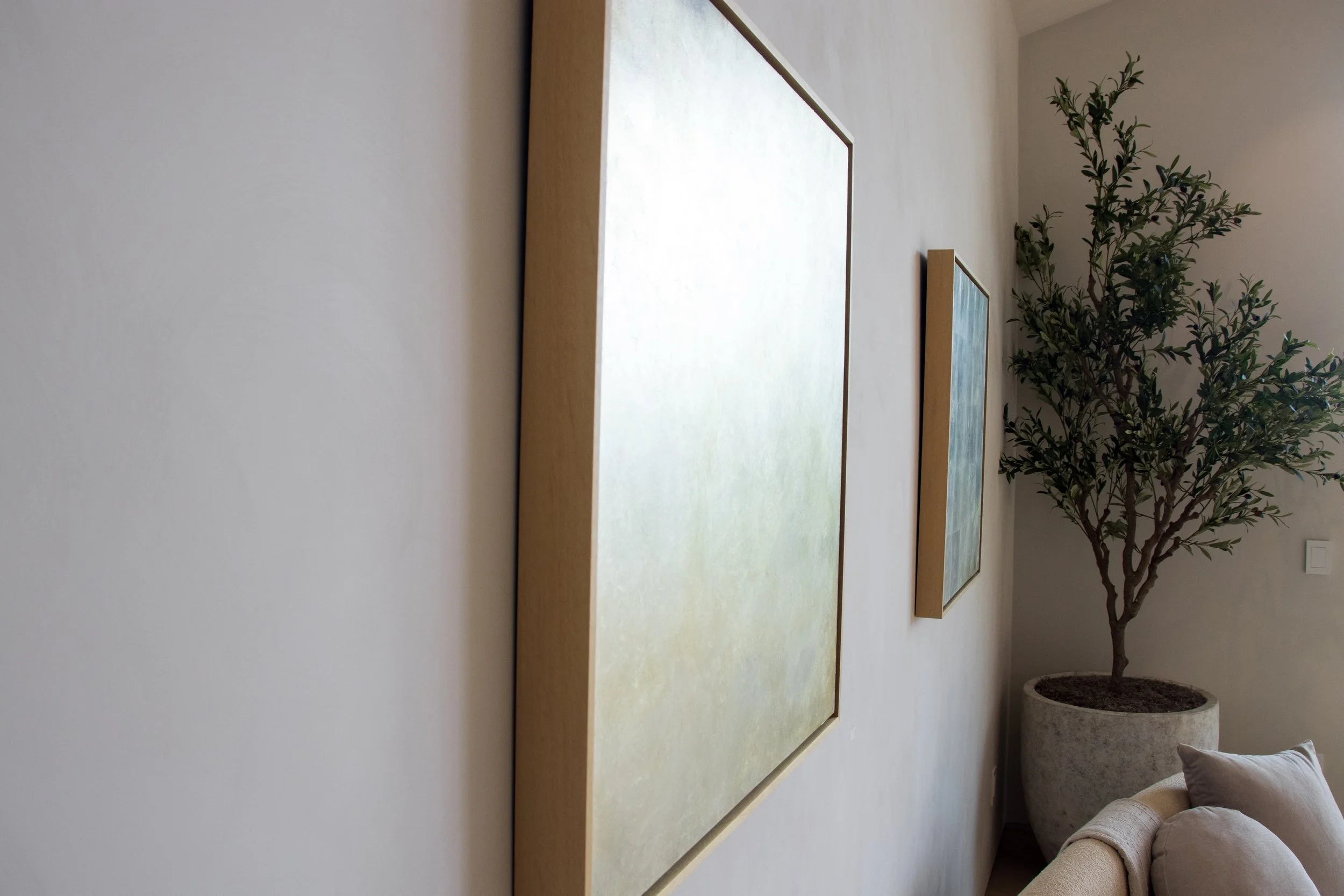 Interior of a room featuring three large framed artworks on a plain wall, a tall potted plant, and a beige sofa with cushions.