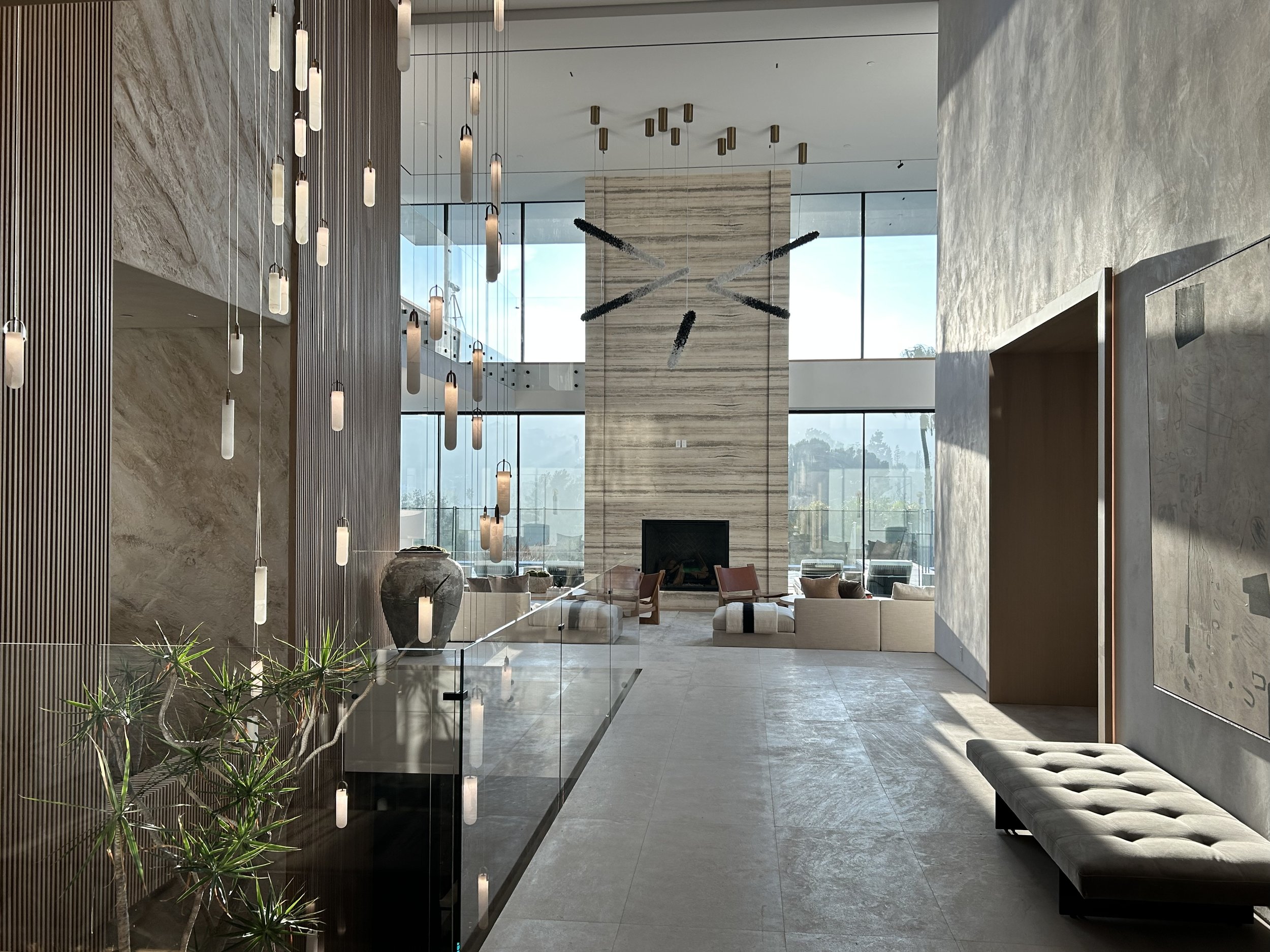 A spacious, modern lobby with large floor-to-ceiling windows, tall hanging light fixtures, a marble fireplace, and a seating area with beige couches and wooden chairs.