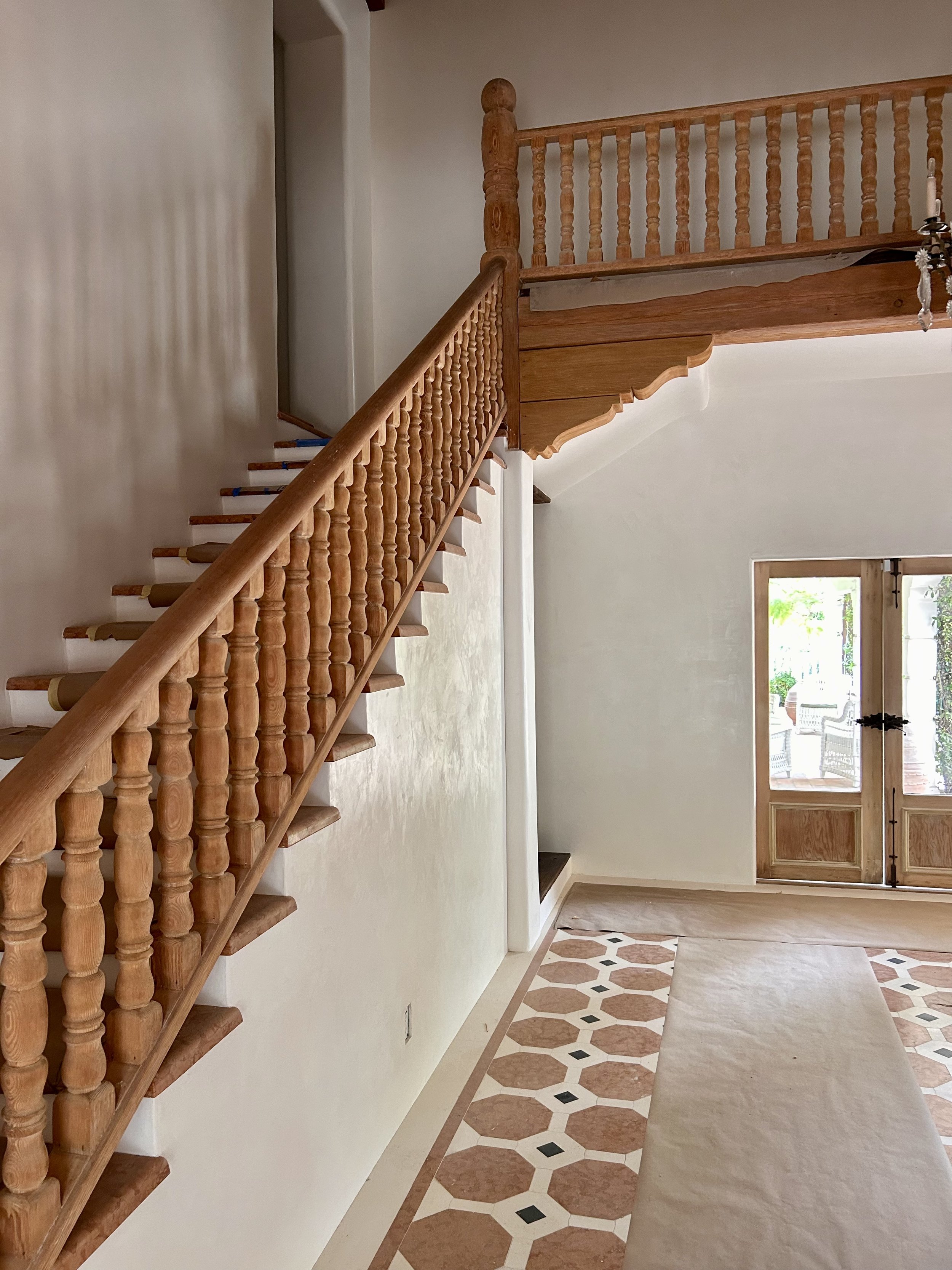 A hallway with a wooden staircase railing on the left, leading to an upper floor. The hallway has a patterned tile floor with a beige runner rug. There is a door with glass panels and a view of the outside, including greenery and a patio with chairs,