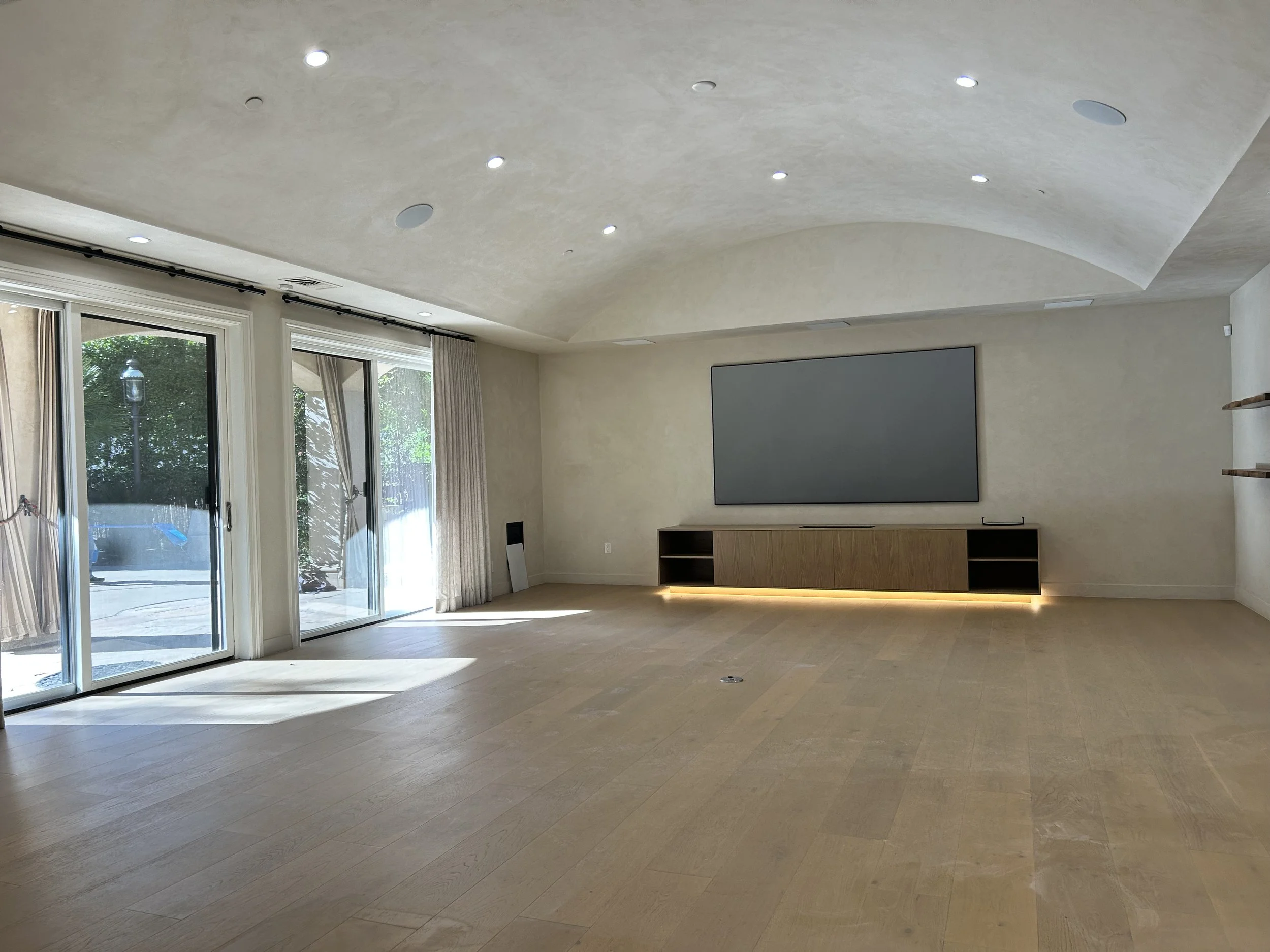 Empty modern living room with large glass sliding doors, beige curtains, and a wall-mounted flat-screen TV, with hardwood flooring and a ceiling with recessed lighting.