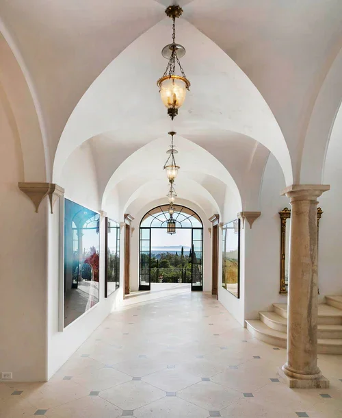 Elegant hallway with white vaulted ceilings, hanging chandeliers, large windows, and a view of greenery and water outside.