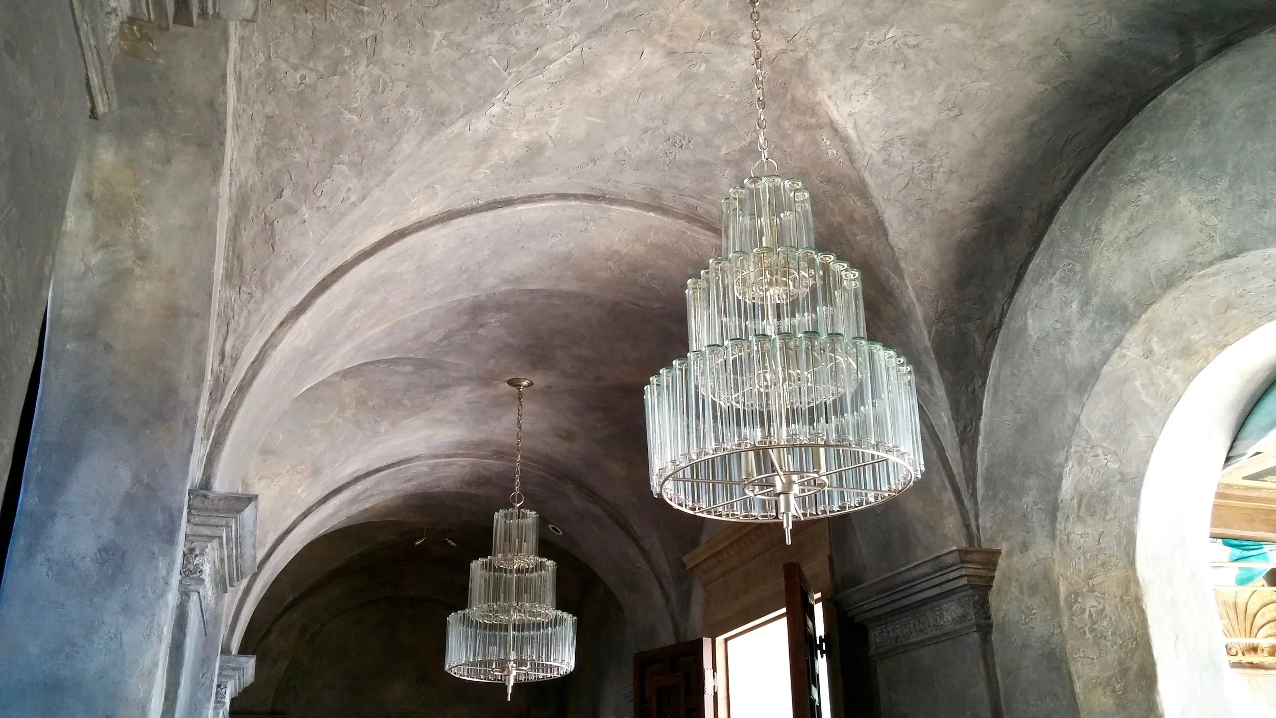 Pendant chandelier with multiple tiers of clear glass tubes hanging from a gold frame, suspended from a vaulted ceiling with a textured stone finish.