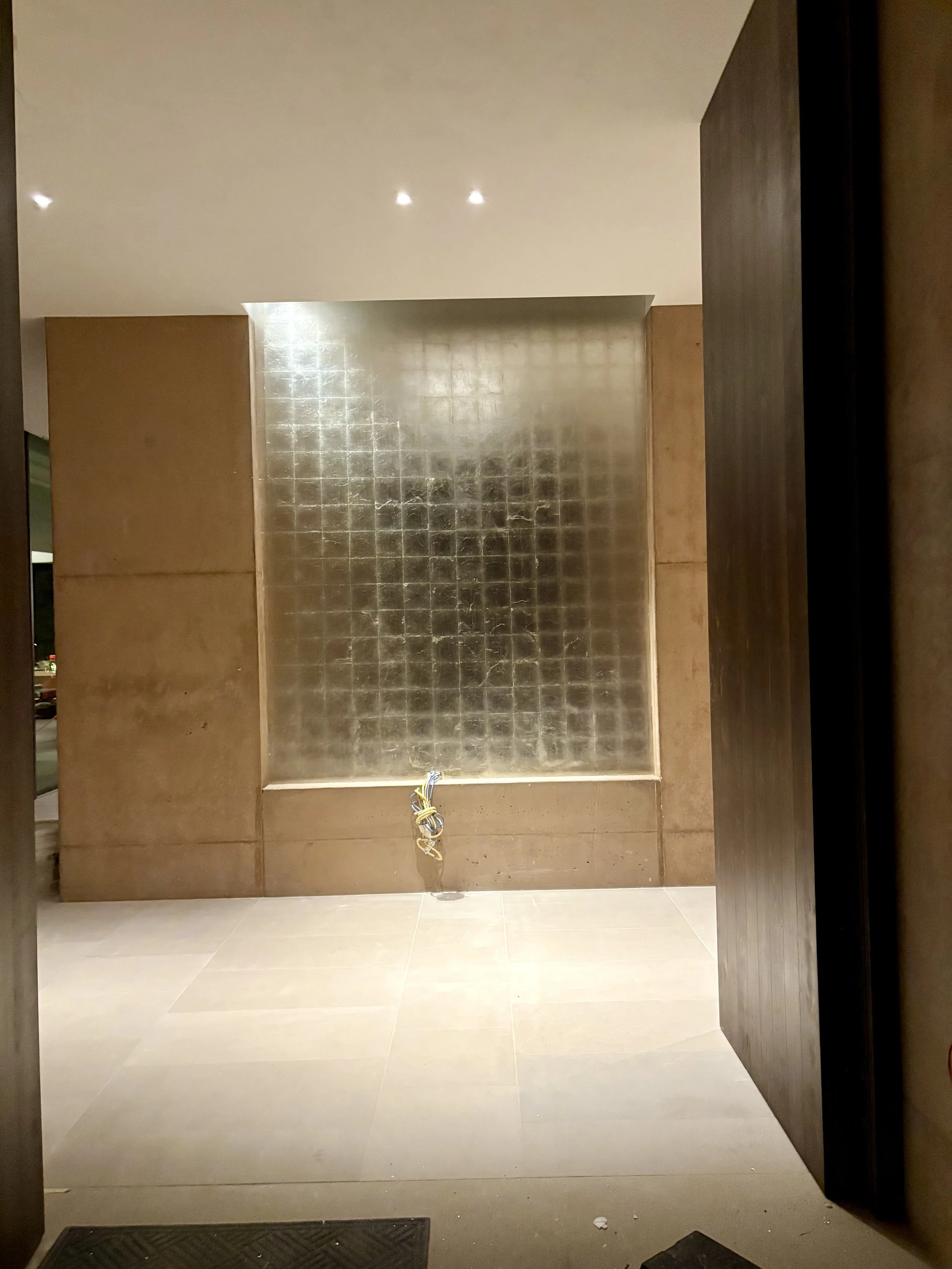 Empty hallway or lobby area with beige tiled floor, beige walls, and a decorative wall feature made of reflective tiles. There are dark wooden panels on either side, and some exposed wires hanging from the center of the wall.