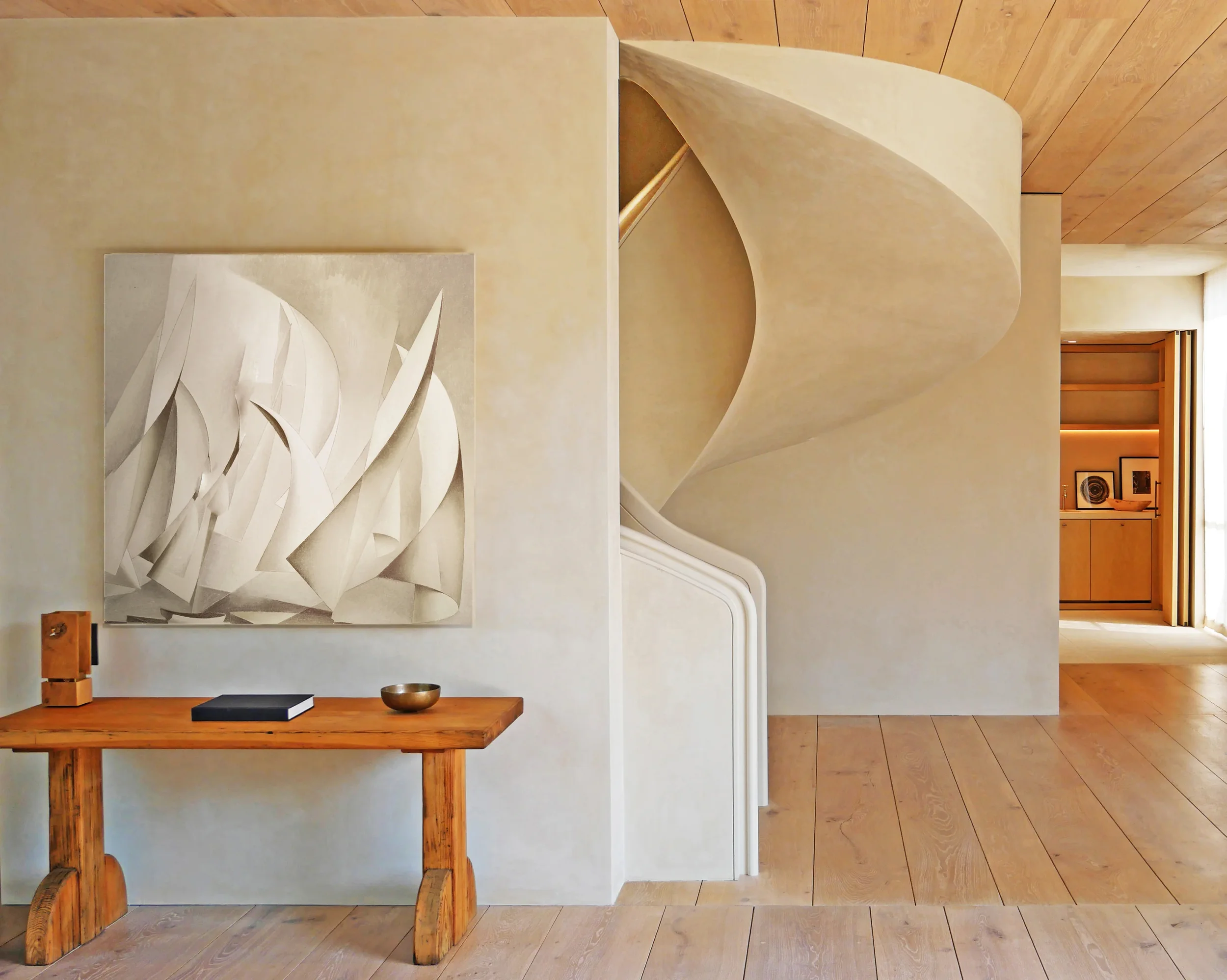 A wooden table with a black book, a small bowl, and a wooden box, against a beige wall with a white abstract sculpture and artwork, in a room with wooden flooring and a doorway leading to another space.