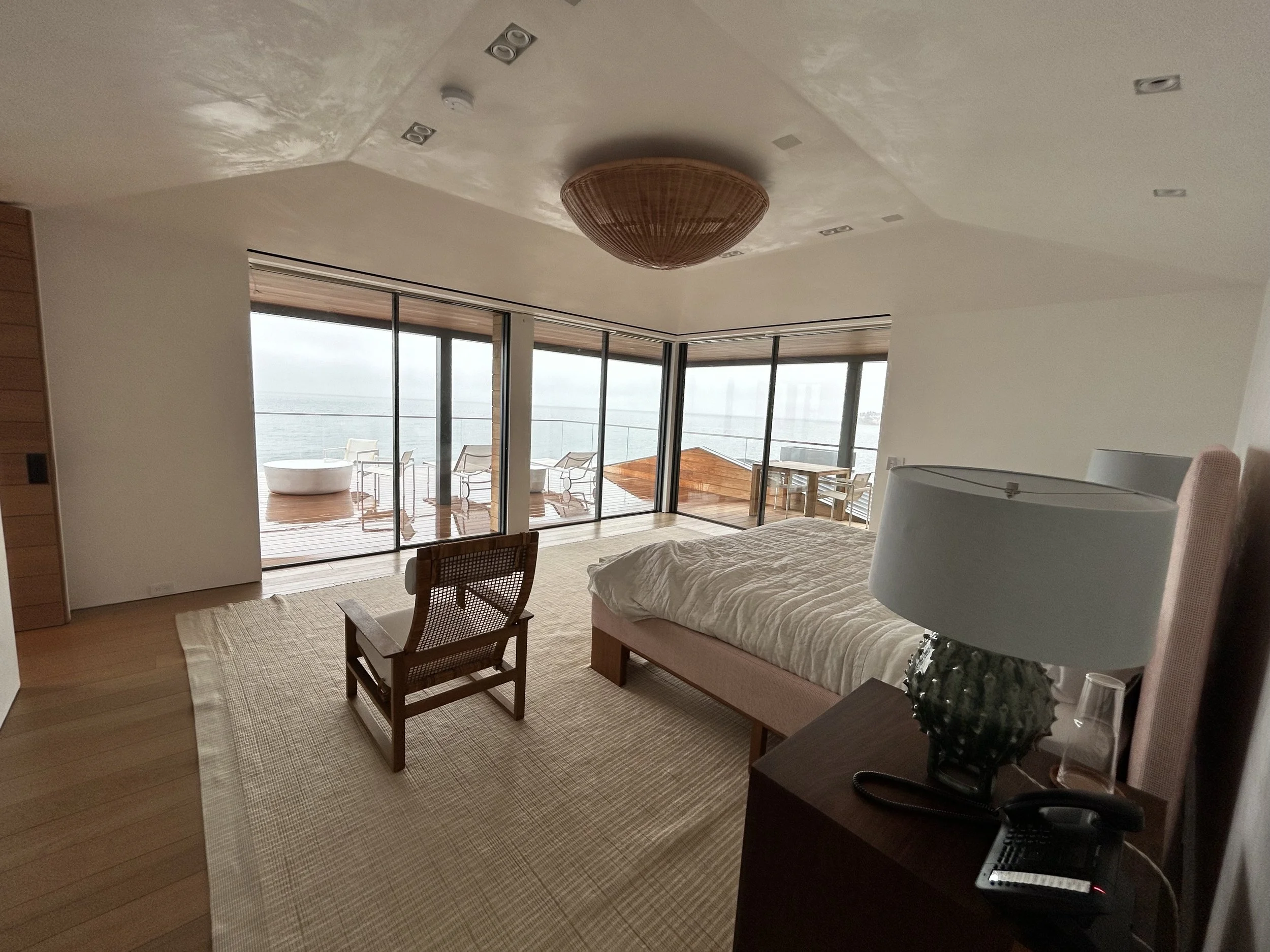 Interior of a modern apartment with glass doors leading to a balcony overlooking the ocean, with outdoor furniture, a bathtub, and a ceiling light fixture.