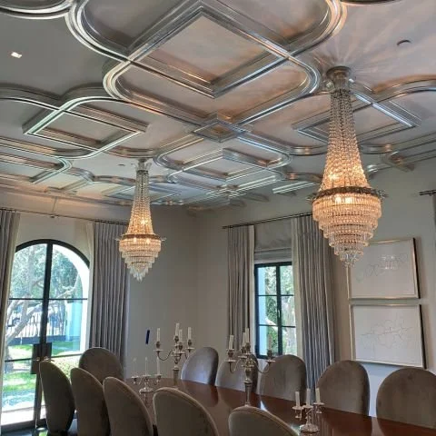 A luxurious dining room with ornate ceiling molding, two large crystal chandeliers, a wooden dining table, upholstered chairs, and large windows with curtains.