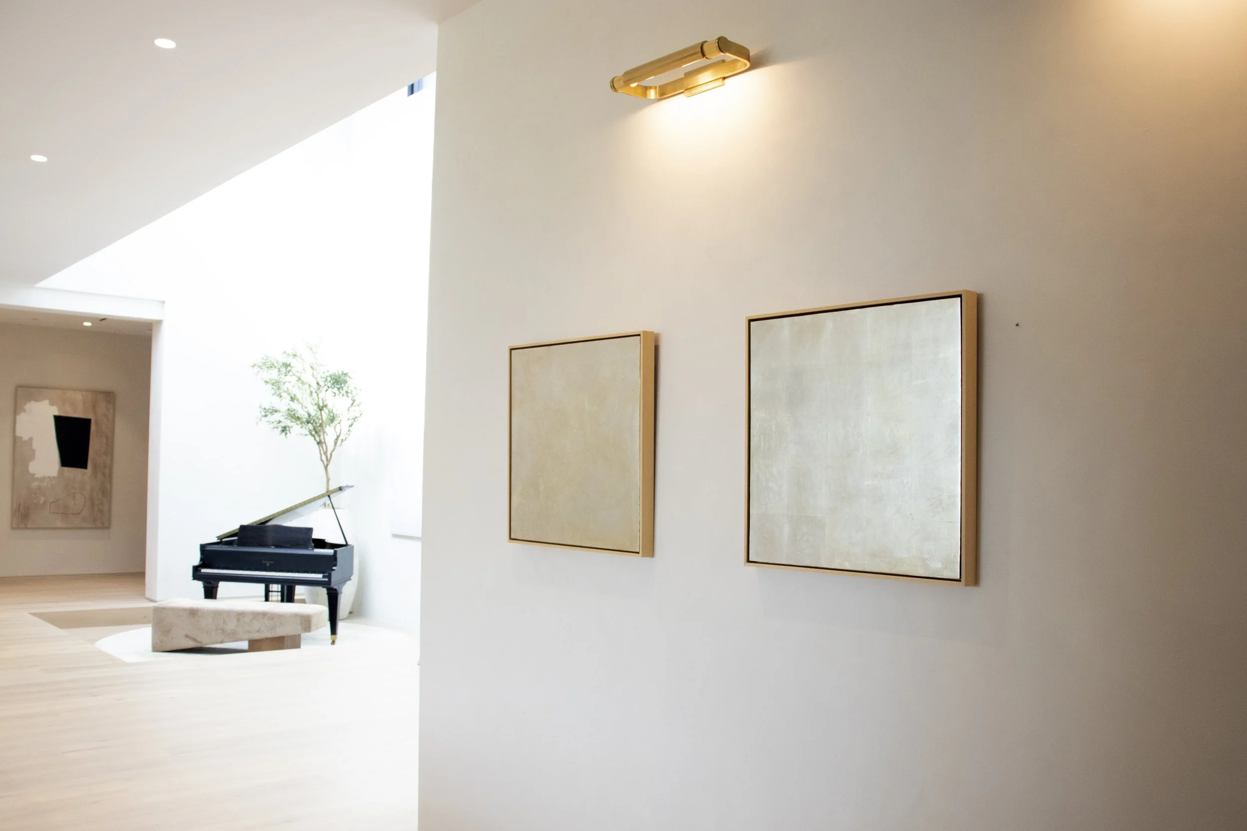 A modern, minimalist interior of an art gallery or living space with white walls and light wooden floors. On the right wall, there are two large, square, beige-colored artworks with simple wooden frames. In the background, a black grand piano is visible next to a small, beige, marble-topped table. There is a tall, leafy indoor plant near the piano, and natural light floods the space through large openings, creating a bright and airy atmosphere.